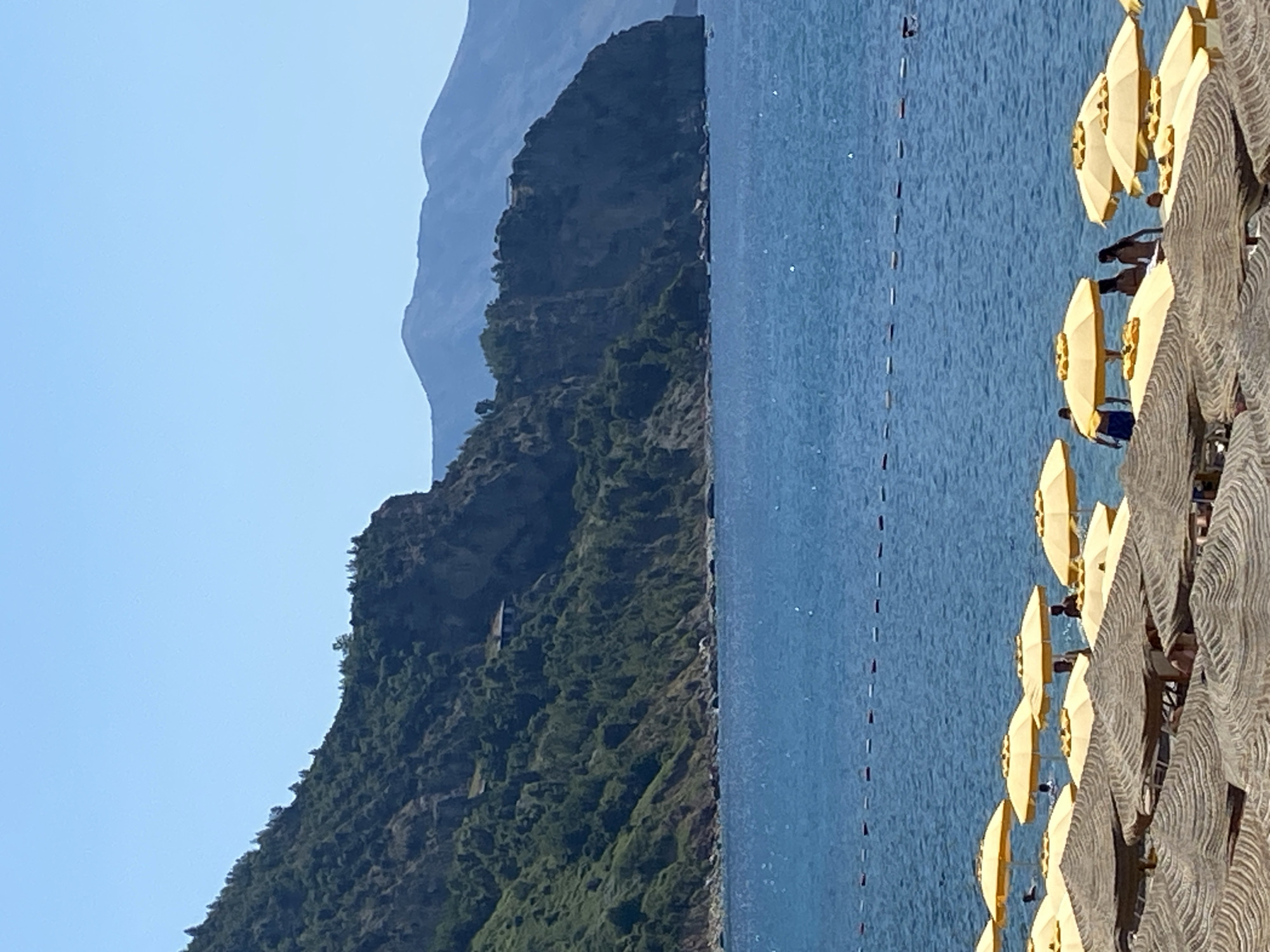 The image depicts a sun-drenched beach scene in Prijevor, Montenegro. The foreground is dominated by rows of thatched and yellow beach umbrellas, casting shadows on the sand. Several people are visible beneath the yellow umbrellas, some appearing to be socializing. In the middle ground, the clear blue Adriatic Sea stretches out, marked by a series of buoys forming a boundary in the water. The background features a dramatic, lush green mountainside that slopes down to the sea, with a few scattered buildings visible on its higher reaches. In the far distance, hazy blue mountains complete the picturesque coastal landscape under a clear, bright sky.