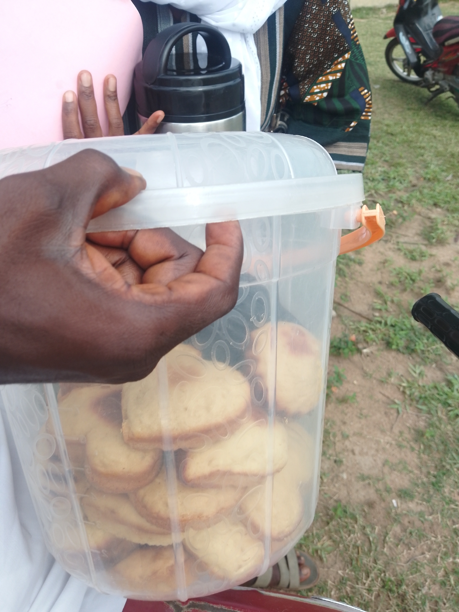 In the city of Serti, Nigeria, a person's hand holds a clear plastic container filled with sliced bread. The container is round and has an orange latch on the side. Behind the container, a pink fabric is visible, along with part of a black and silver thermos. The person is wearing a colorful patterned shirt. The background shows a grassy area with some dirt patches and a red motorcycle parked in the distance. The overall lighting suggests it is daytime.
