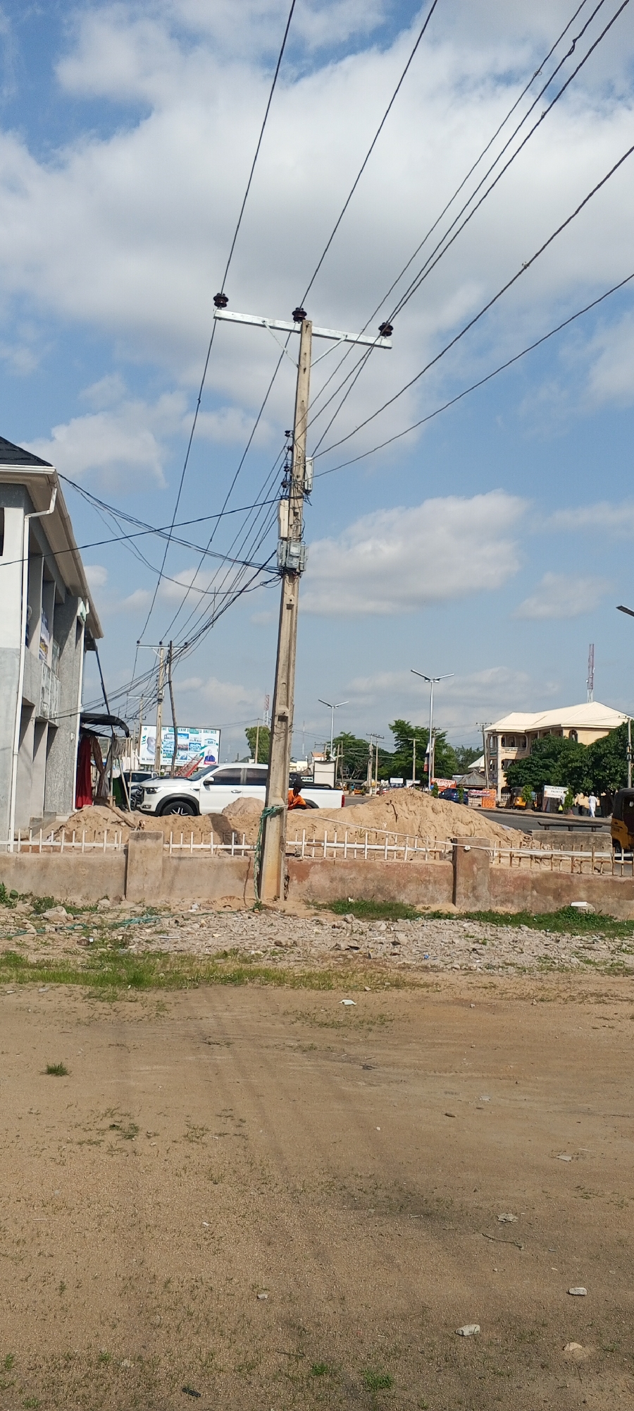 The image depicts a street scene in Jalingo, Nigeria, characterized by a prominent utility pole with numerous electrical wires crisscrossing the frame. In the foreground, a dusty, unpaved area with sparse grass suggests a construction or undeveloped plot. A low concrete barrier, topped with white posts, separates this area from the street. Behind the barrier, a large mound of sand is visible, indicating ongoing construction or roadwork. A white pickup truck is parked near the sand mound.
To the left of the utility pole, a portion of a modern building with a grey facade and white trim is visible. Across the street, several other buildings can be seen, including one with a beige exterior and multiple floors, and another with a red roof. Streetlights are scattered along the roadside. Several vehicles, including what appear to be tricycles and other cars, are present on the street. The sky is partly cloudy, with scattered white clouds against a blue backdrop. The overall impression is a functional urban environment with elements of ongoing development.
