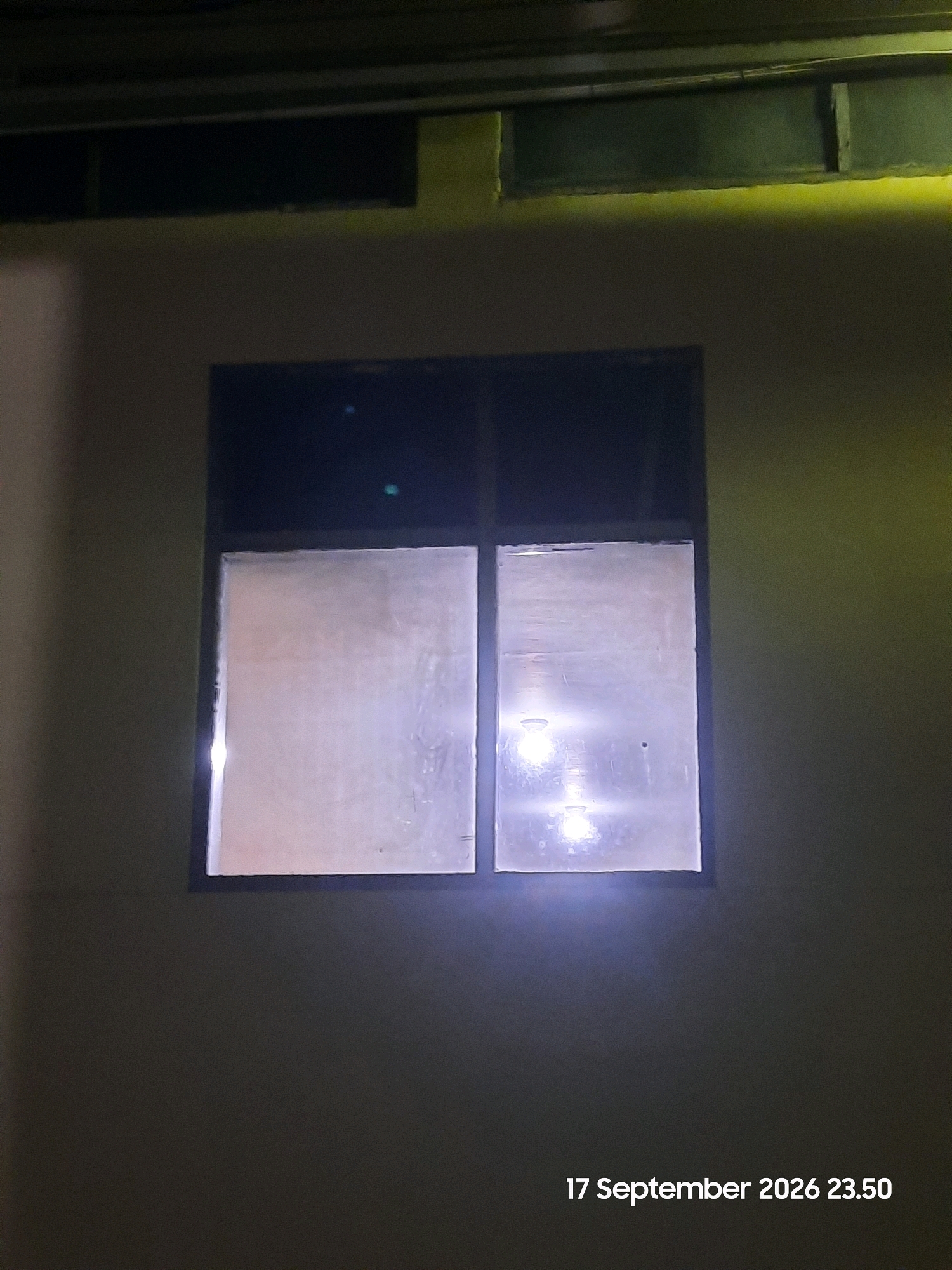 A nighttime exterior shot shows a building wall with a large, darkened window divided into two panes. The window panes are covered with a translucent material that diffuses the light from within. Two bright, circular light sources are visible within the right pane, suggesting interior lighting. The wall appears to be constructed of concrete and is dimly lit by an unseen light source, casting a yellow hue in the upper portion of the image. The bottom right corner of the image displays a timestamp: "17 September 2026 23.50". There are no people or active subjects depicted in the image. The location is stated to be Gending, Indonesia.