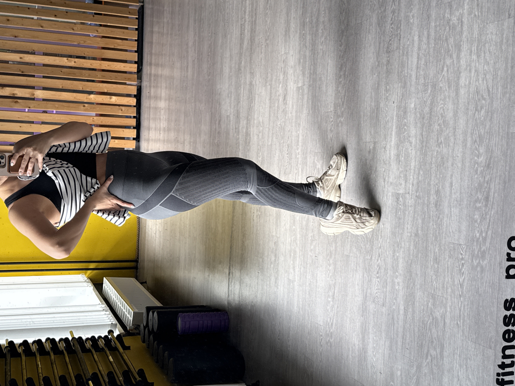 A person wearing a black sports bra, a striped top, grey leggings, and beige sneakers stands on a grey wooden floor with "fitness pro" printed on it. The person is holding a smartphone to take a mirror selfie. The background includes a wall with wooden slats, a yellow wall, fitness equipment (dumbbells, kettlebells), and a white radiator. The location is inside a fitness studio in Izhevsk, Russia.
