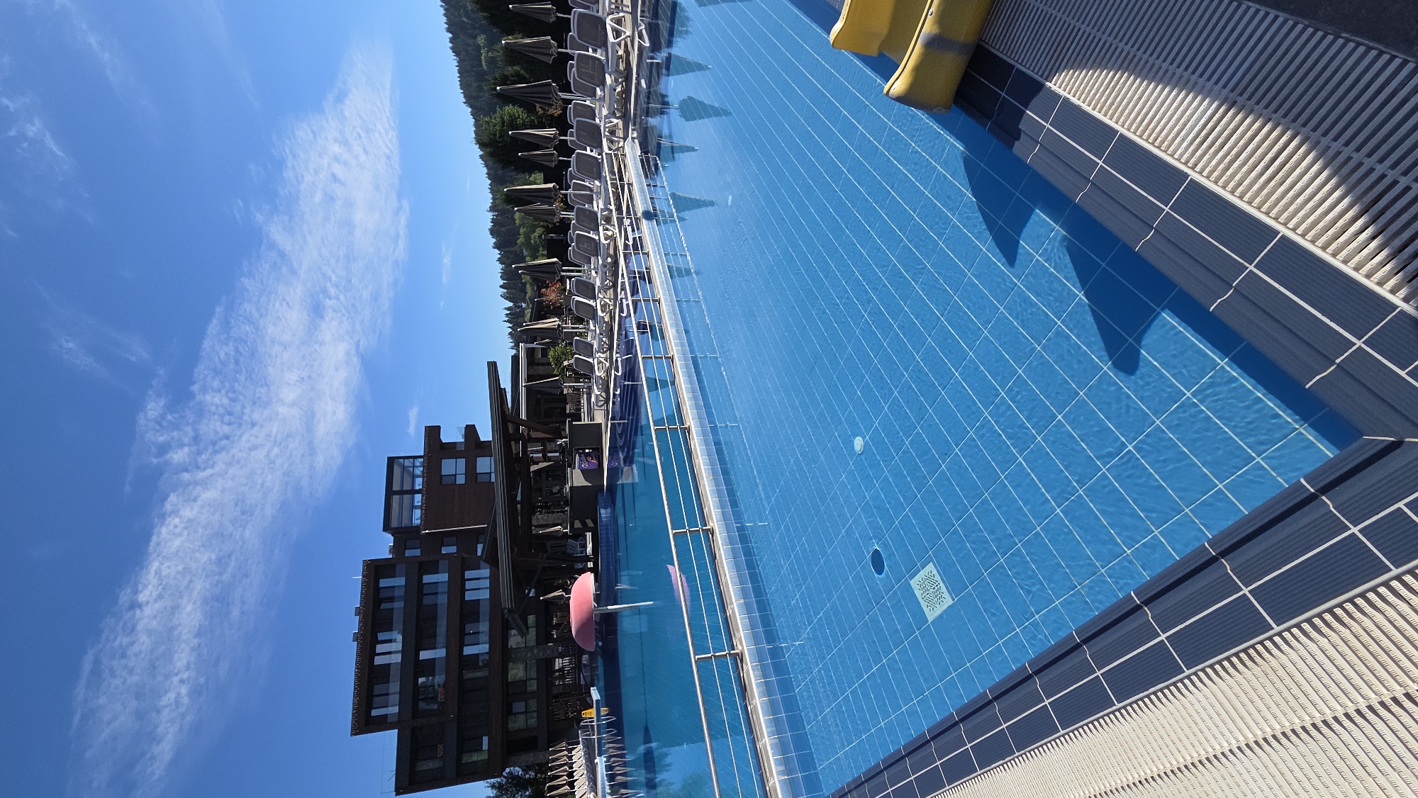 A bright, sunny day is depicted with a wide shot of a blue-tiled swimming pool in the foreground. The pool is empty of people. In the background, a modern, multi-story wooden building with many windows stands against a clear blue sky dotted with wispy white clouds. Several rows of empty lounge chairs and closed umbrellas are lined up on the pool deck. To the far left, part of a pink inflatable object is visible in the shallow end of the pool. To the far right, two yellow sections of a slide are partially visible at the edge of the pool. The surrounding environment includes lush green trees and hills in the distance. The overall scene suggests a resort or hotel setting.