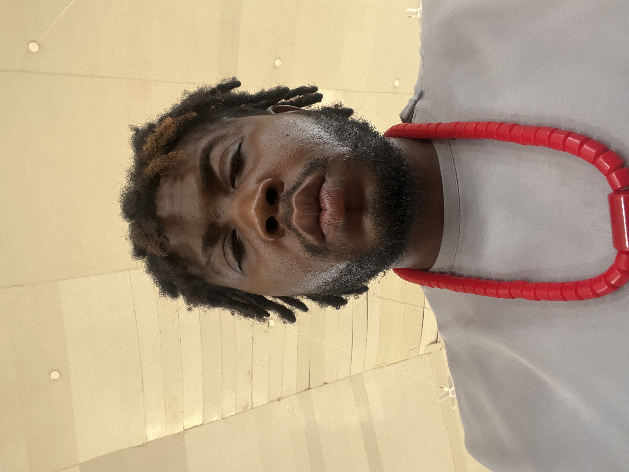 A close-up, waist-up shot shows a man with dark skin, dreadlocks with brown highlights, and a beard. He is wearing a gray shirt and a bright red necklace with large, cylindrical beads. The man is looking directly at the camera. The background is a ceiling with beige panels and recessed lighting. The lighting suggests an indoor location, possibly a commercial building or an event space. There is no clear indication of the specific location within Barakin Akawo, Nigeria, beyond the provided context.