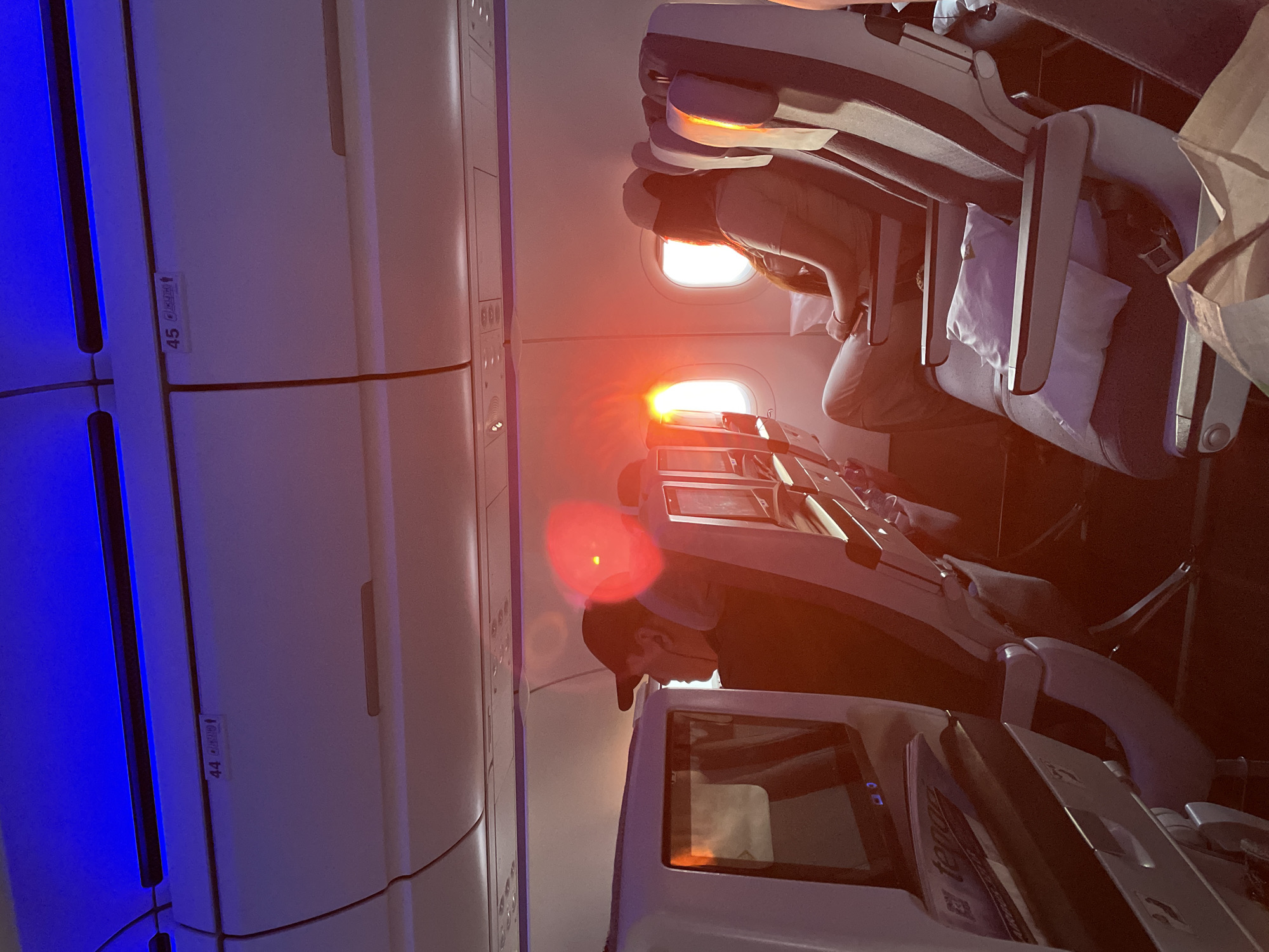 The image depicts the interior of an airplane cabin during sunset. The setting sun casts a warm, orange glow through the windows, illuminating the aisle and the seats. Passengers are seated, with one individual in the foreground wearing a baseball cap and listening to headphones. Another passenger is visible further down the aisle, facing the window. Overhead compartments and seatback entertainment screens are visible. The lighting suggests it is dusk, and the blue ambient lighting at the top of the cabin adds to the atmospheric quality of the scene. There is no indication that this image is located in Beykoz, Turkey.