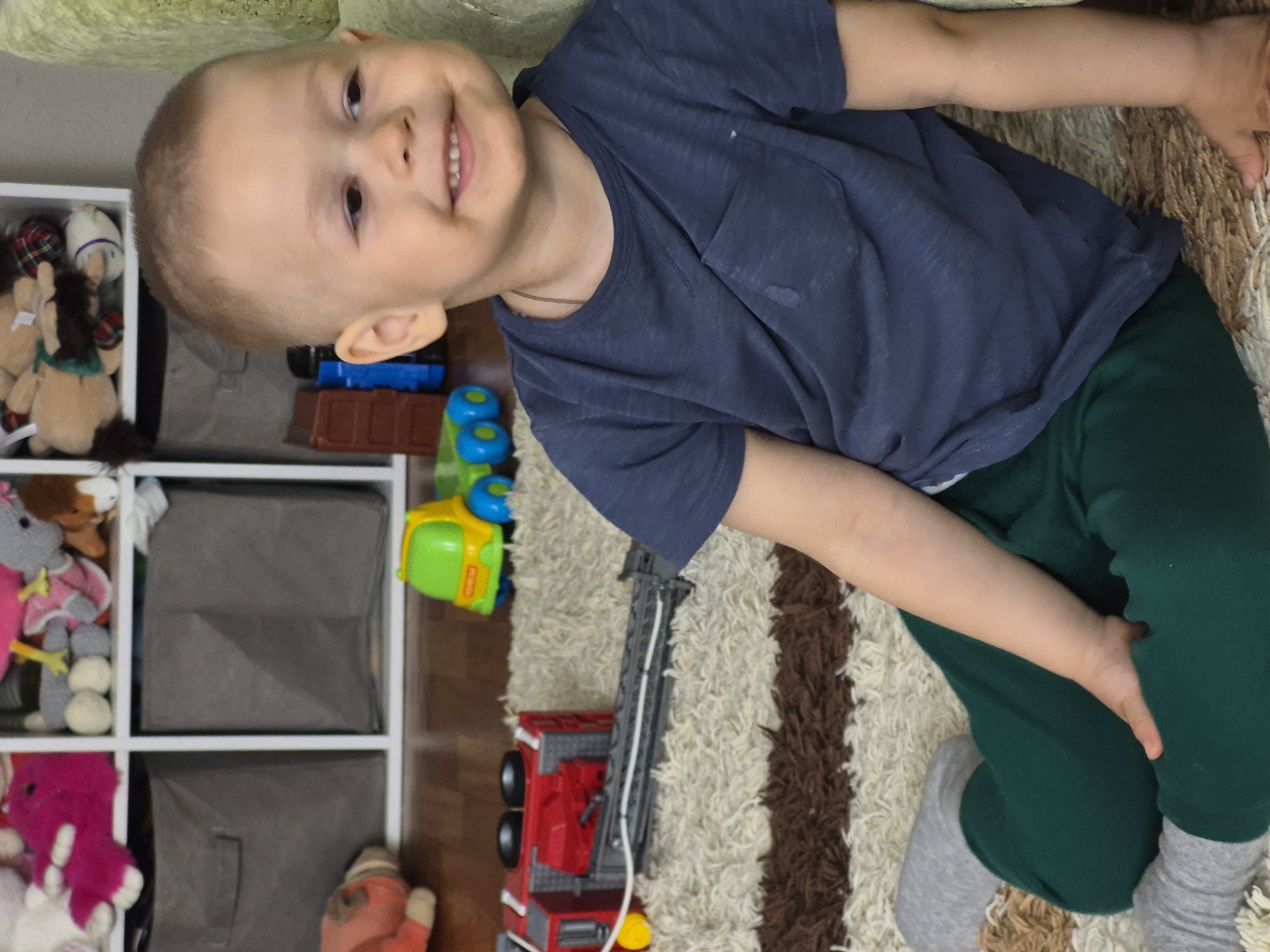 A young child, likely between 2 and 4 years old, is positioned in the frame. The child has a fair complexion, a close-cropped haircut, and is smiling. The child is wearing a dark blue, short-sleeved t-shirt and green pants, with grey socks visible on their feet. The child is lying on a patterned rug with cream and brown stripes.
Next to the child is a white shelving unit filled with stuffed animals and grey storage bins. A variety of toys, including a green and yellow toy truck and a red toy fire truck with an extending ladder, are scattered on the rug. The image is taken indoors. The location is not discernible from the image, so it cannot be determined if it is in Tobolsk, Russia.