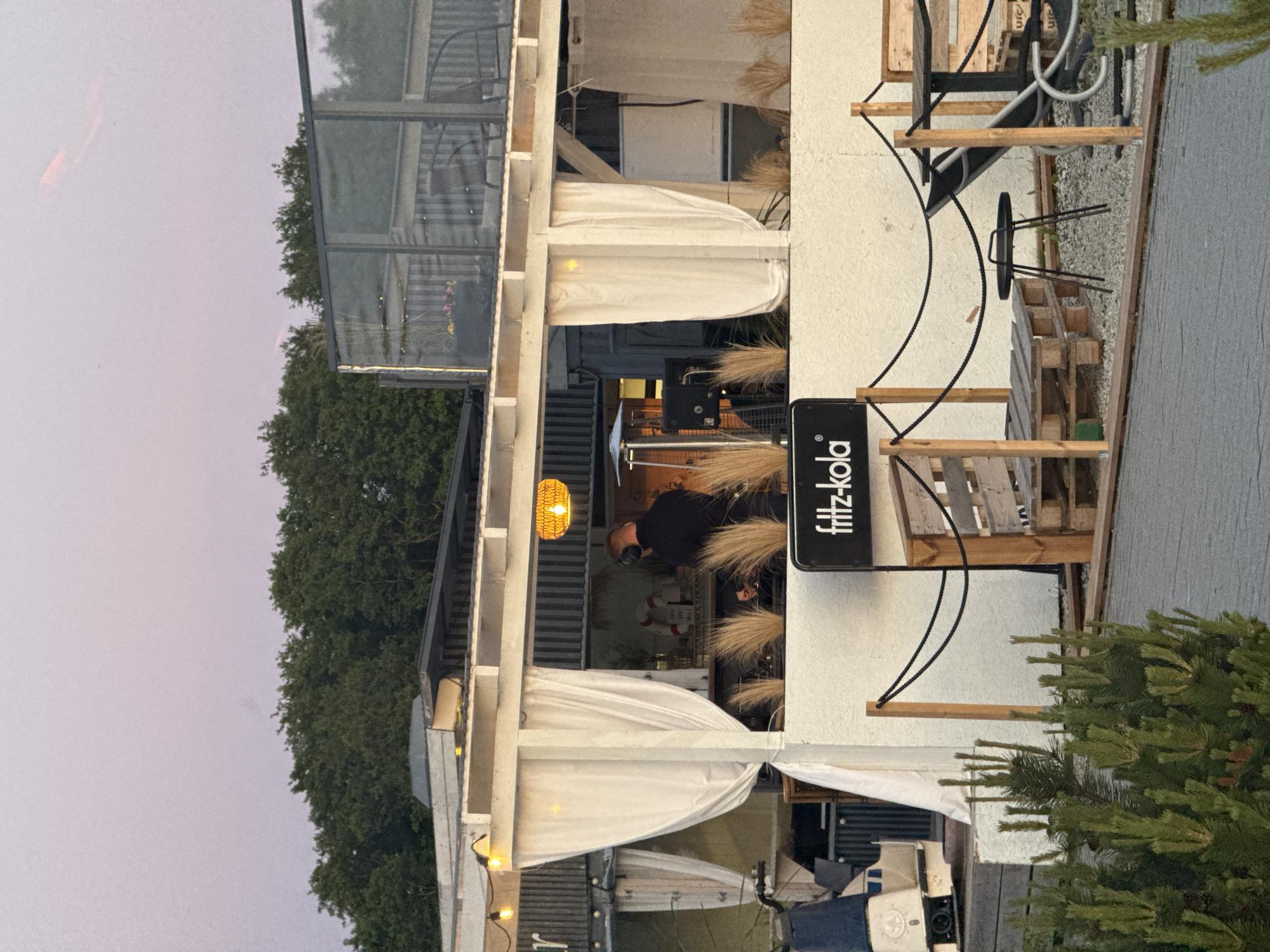 The image captures an outdoor seating area at a bar or restaurant in Tallinn, Estonia, likely during the evening given the soft light and the pinkish hue of the sky. In the background, a structure that appears to be a shipping container converted into a bar is visible, draped with white curtains. A person wearing headphones and a dark shirt is working behind the counter, possibly a DJ or bartender. On the counter, there are bottles, a life preserver, and what looks like DJ equipment. In the foreground, there are several outdoor furniture pieces, including wooden pallet benches, a small black metal table, and chairs. A black sign with white lettering that reads "fritz-kola" is prominently displayed. The ground is covered with gravel and a paved walkway. Green bushes are in the lower left corner of the image. The overall atmosphere suggests a relaxed, casual setting by the waterfront, possibly a beach bar.