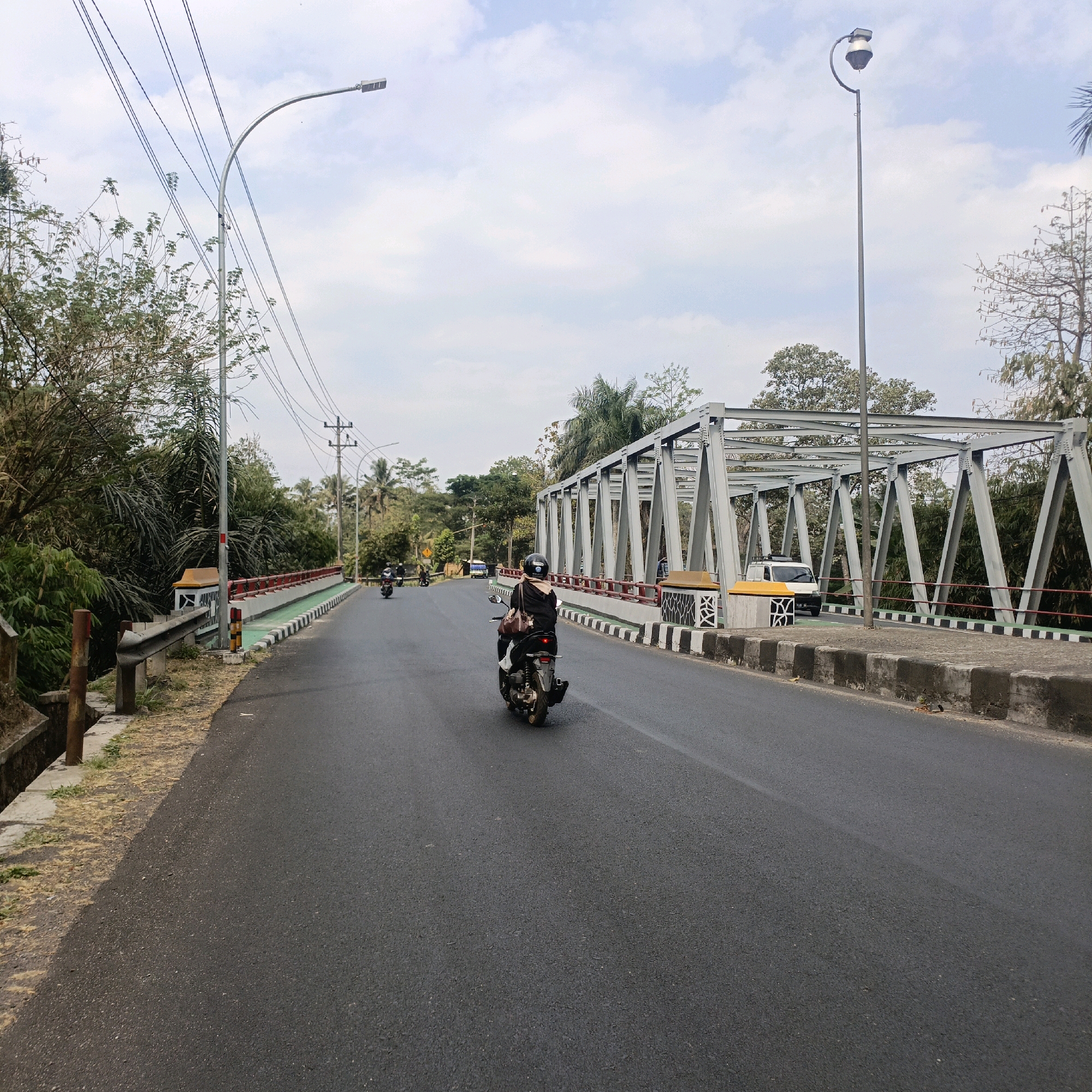 A street scene in Salatiga, Indonesia, features a paved road with a metal truss bridge on the right. The road has a dashed white line marking the lane. A person on a motorcycle is in the foreground, riding away from the camera. Several other motorcycles are visible further down the road. On the left side of the road, there is a concrete barrier with red and white markings, and lush green trees and vegetation. A tall street lamp with a curved arm extends into the sky, with power lines running across the frame. The sky is partly cloudy. The bridge has a grey metal structure with diagonal supports. A white van is parked on the bridge. The environment appears to be a mix of urban infrastructure and natural foliage.