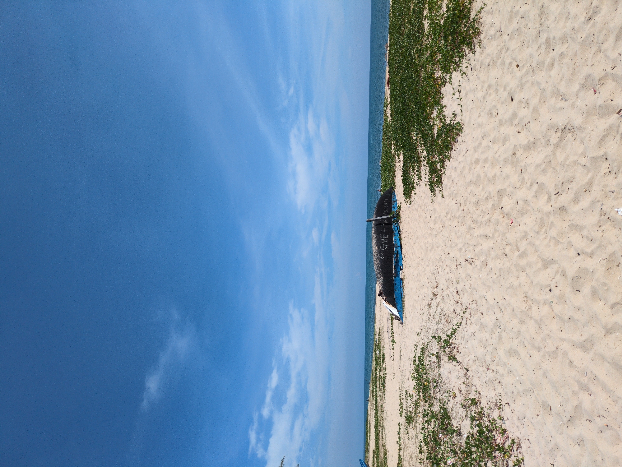 The image depicts a serene beach scene in Thuan An Ward, Vietnam. The foreground is dominated by a sandy beach dotted with scattered green vegetation. In the mid-ground, an overturned blue and black boat rests on the sand. The boat has some writing on its side, appearing to be "GHE + MA" followed by numbers. Beyond the beach, the calm blue ocean stretches to the horizon, meeting a clear blue sky with a few wispy white clouds. The overall impression is one of peacefulness and natural beauty, with no people visible in the frame.