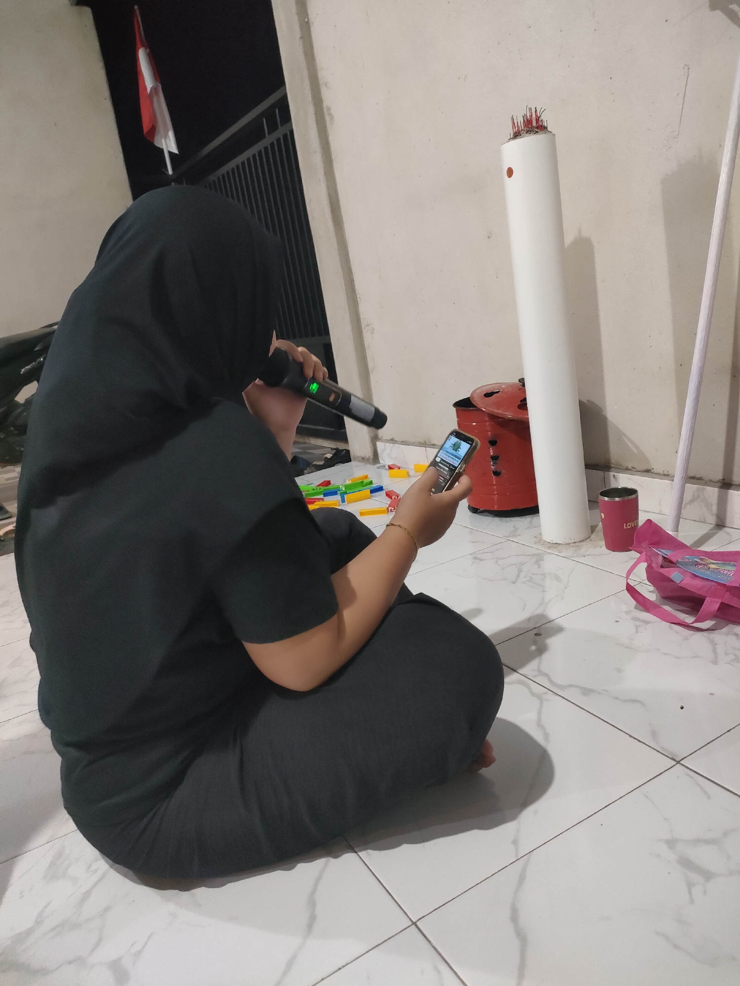 A person wearing a black hijab and black clothing sits on a tiled floor, holding a microphone to their mouth and a smartphone in their right hand. The person appears to be singing or speaking into the microphone while looking at the phone. Colorful building blocks are scattered on the floor to the left of the person. In the background, a red container, a white cylindrical object, and a pink bag are visible. A white pole stands to the right of the white cylinder. An Indonesian flag is displayed on a pole at the top left of the image. The setting appears to be an outdoor area, possibly a porch or patio, with a wall in the background.
