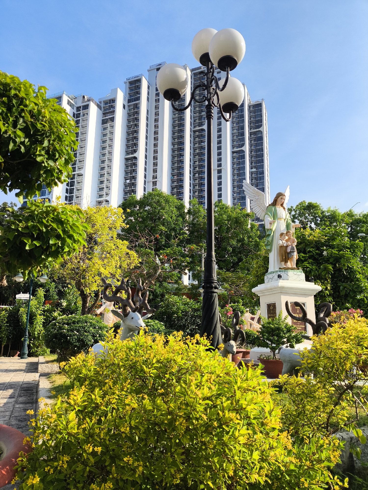 In the city of Bac Nha Trang Ward, Vietnam, a modern white high-rise building with many windows stands tall against a clear blue sky. In the foreground, a garden features lush green trees and bushes, some adorned with small yellow flowers. A multi-headed lamppost with white globes is positioned in front of the building. Among the foliage, there are several statues, including a prominent angel statue holding two children, a white statue of a goat with large horns, and other decorative figures. A paved walkway is visible to the left of the garden. The scene captures a blend of urban architecture and natural elements, enhanced by religious iconography.