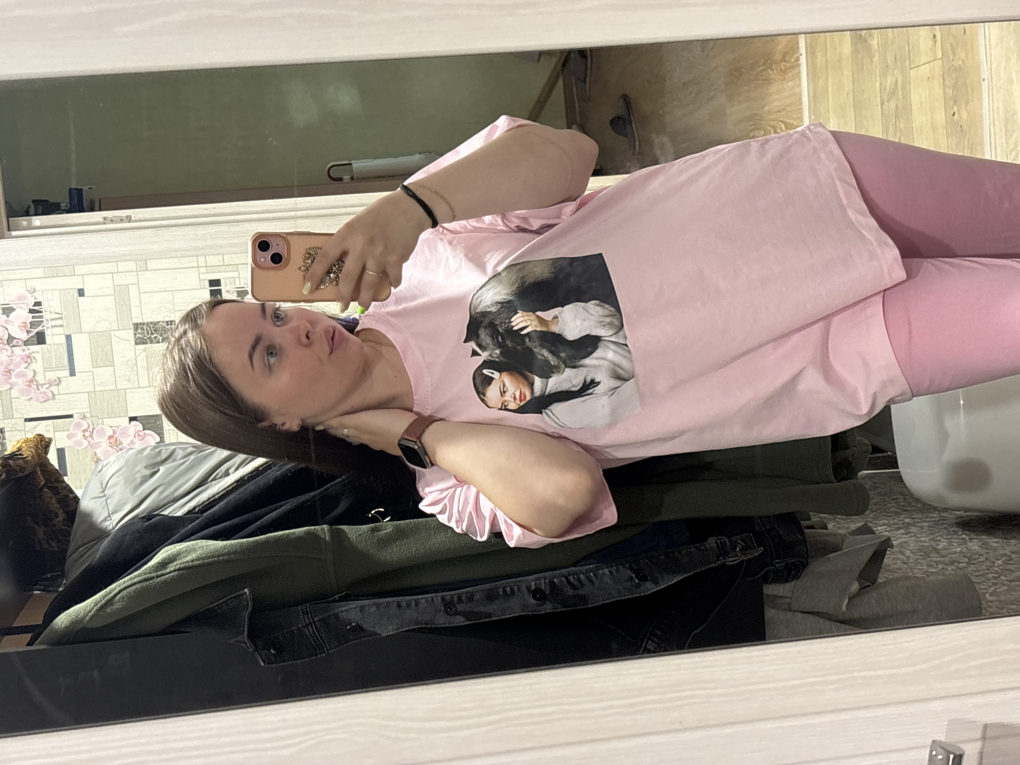 A woman is seen in a mirror taking a selfie with a pink smartphone. She is wearing pink pants and a pink t-shirt with a black and white graphic. The background consists of a tiled wall with floral decor, household items on a shelf, and a wooden floor. In the foreground, there is a pile of clothing including a green jacket and a black denim jacket. The location is potentially Izhevsk, Russia, based on visual cues.