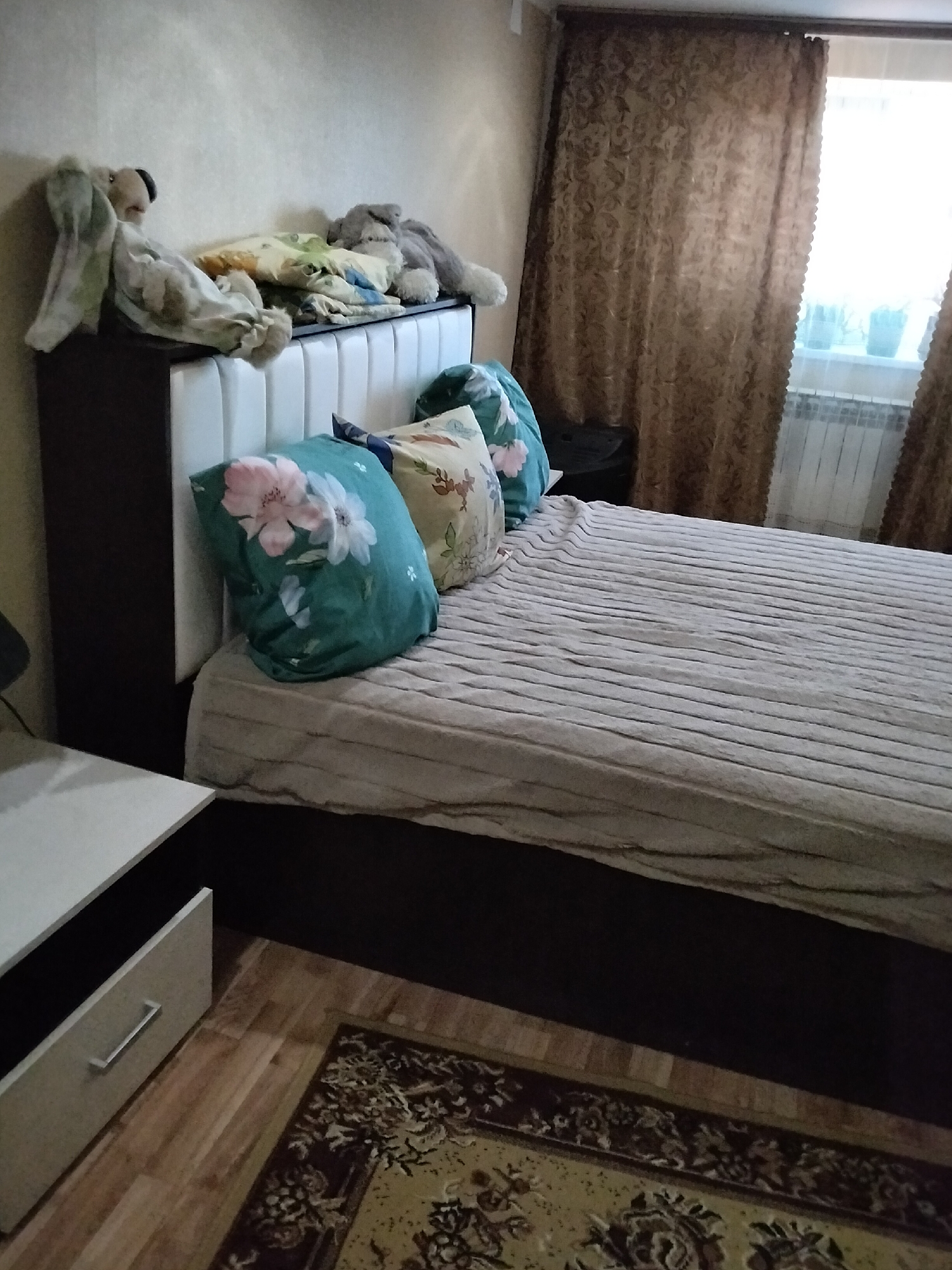 The image shows a bedroom with a bed, two nightstands, a rug, and a window with curtains. The bed is made with a beige blanket and decorated with two pillows, one with a floral pattern and the other with a floral pattern. On the headboard of the bed, there are several stuffed animals, including two bunnies and a dog. To the left of the bed is a white nightstand with a drawer. To the right of the bed, a dark-colored nightstand is partially visible. The floor is covered with a patterned rug and wooden planks. The window, covered by brown patterned curtains, is to the right of the bed. The lighting in the room appears to be natural, coming from the window. The overall impression is a lived-in bedroom. The location is stated to be Nizhnedonskoy, Russia.
