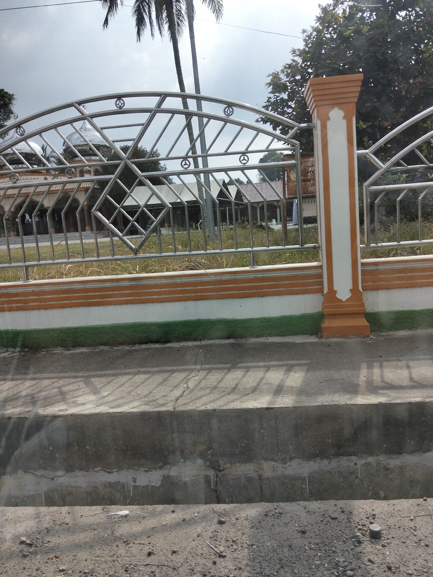 The image shows a view of a mosque through a metal fence in an outdoor setting. The mosque has a silver dome and is partially obscured by the fence, which features a decorative pattern of diamond shapes. The fence is supported by concrete pillars, one of which is white with orange trim and decorative accents. Beyond the fence, there is dry grass and some trees. In the foreground, there is a concrete pathway or curb, with shadows cast by the fence. The sky is overcast. The image appears to be taken from a street or sidewalk, with a view looking towards the mosque and its surrounding area. There are no people clearly visible in the frame. The overall scene suggests a public space, likely in a warm climate given the presence of palm trees. The context of "Unknown, Indonesia" suggests a location within Indonesia.