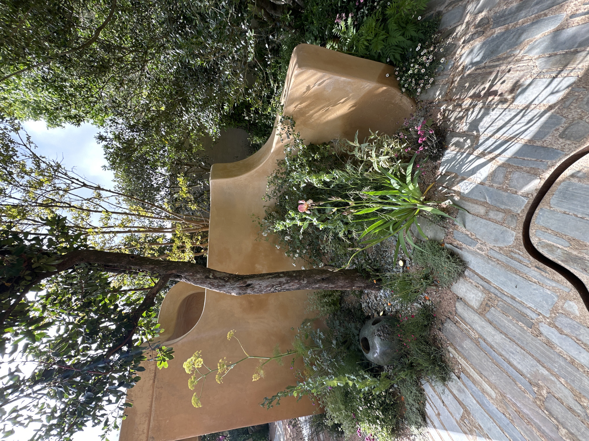 The image depicts a garden scene, possibly in London, United Kingdom. It features a curved, light-brown concrete structure overgrown with various plants and flowers, with a small tree growing alongside it. The ground is covered with gray stone paving, with additional vegetation spilling onto the walkway. A decorative gray urn sits near the base of the structure. The scene is partially shaded by overhead trees. The angle of the shot suggests it was taken from a low perspective.