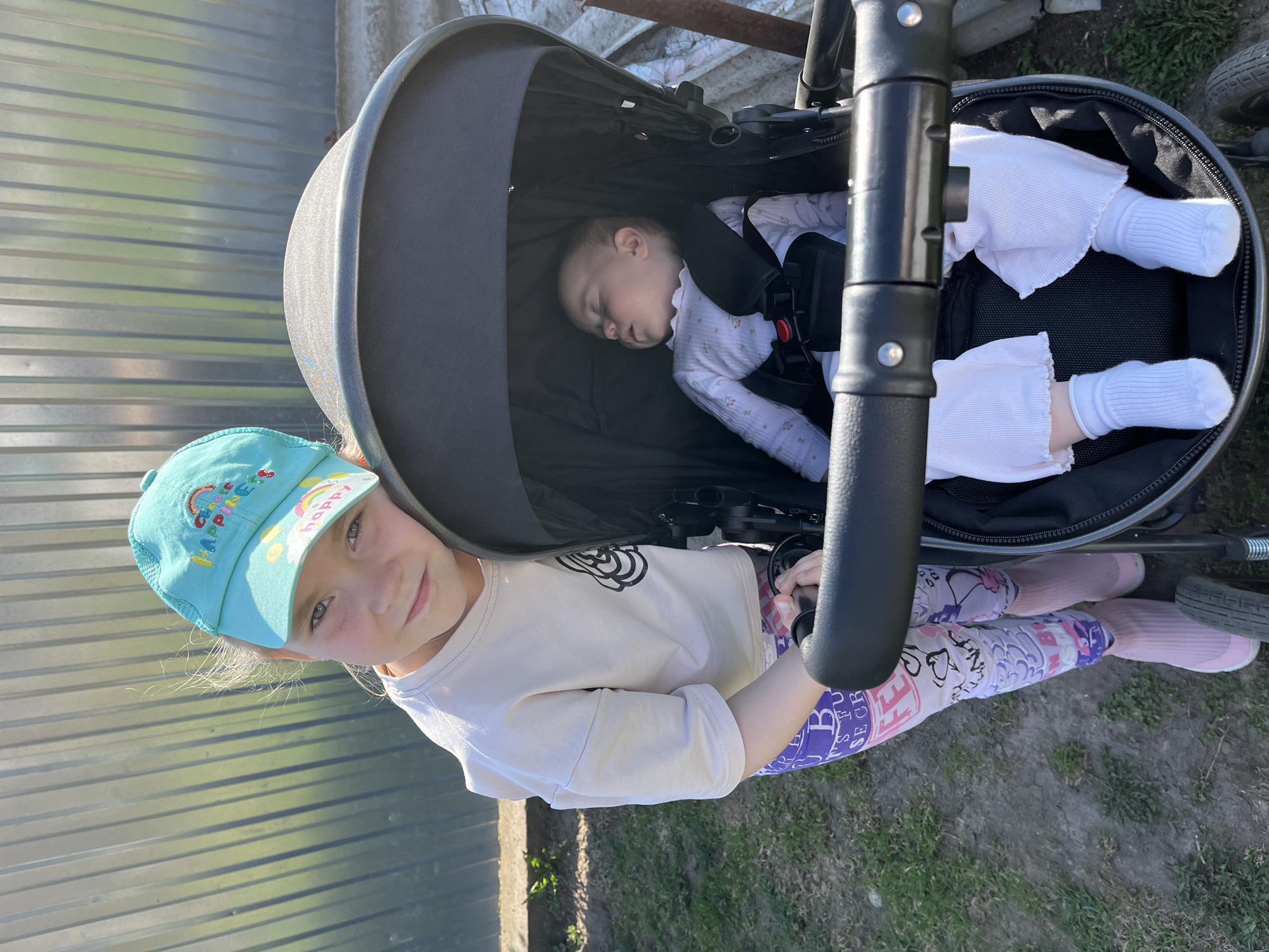 A young girl with a blue cap stands next to a baby stroller, where a baby is sleeping. The girl is wearing a light-colored top and patterned leggings, and the baby is dressed in white. The stroller is black and has a safety harness on the baby. The scene is outdoors, with a metal fence behind the girl and grass visible near the stroller. The location is in Chaikovsky, Russia.