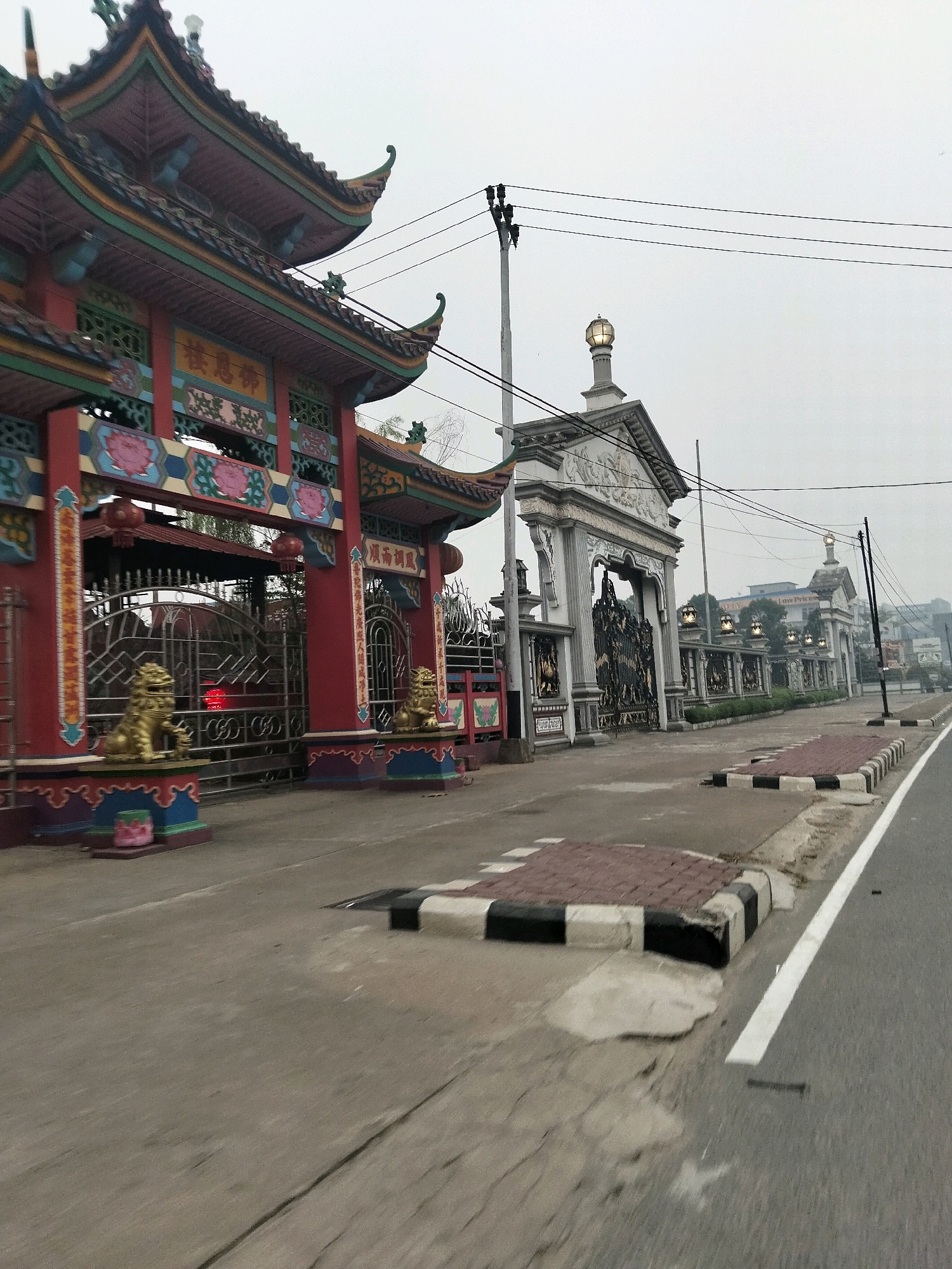 The image depicts an exterior view of a temple gate in Sungai Raya, Indonesia, characterized by its elaborate, ornate design. The gate itself is painted in vibrant red and adorned with intricate carvings and decorative elements, including traditional Chinese-style roofs and golden lion statues flanking the entrance. Beyond the red gate, a second, more austere grey gate with large, ornate metalwork is visible, leading to a long, colonnaded structure with multiple lamp posts. The scene is situated alongside a paved road, with a concrete sidewalk and a traffic lane marked by white lines. The sky is overcast, casting a diffused light over the landscape. The overall impression is one of a grand and historically significant entrance to a place of worship.