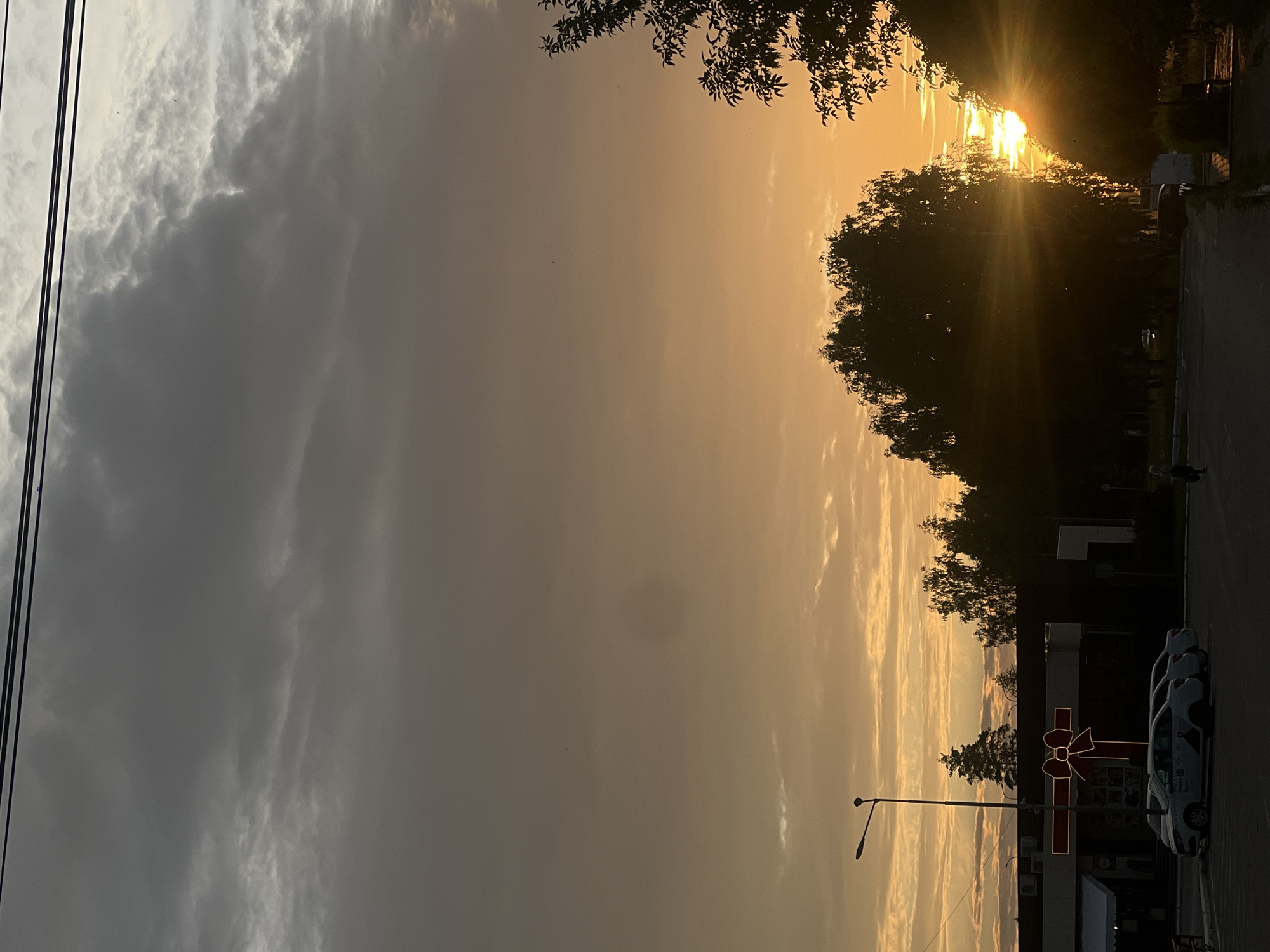 The image depicts a sunset scene in Chaikovsky, Russia, likely during the evening. In the foreground, a street is visible with several cars parked on the right side. A few individuals are present on the sidewalk, one walking. The street is lined with trees, some of which are silhouetted against the bright orange sky. The sky itself is filled with dramatic clouds, transitioning from dark gray at the top to a warm, golden hue closer to the horizon where the sun is setting. The sun's rays are breaking through the trees, creating a lens flare effect. In the background, a building with a large illuminated red bow decoration is visible, suggesting it might be a commercial establishment. A street lamp stands prominently near the building. The overall atmosphere is serene, with the soft light of the sunset illuminating the scene.