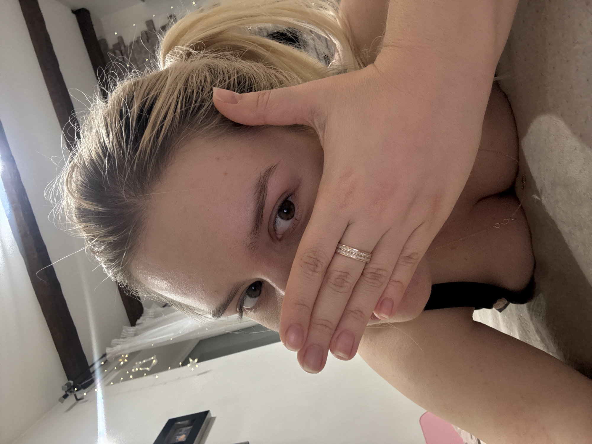 The media file shows a close-up, overhead shot of a person lying down, with their head tilted. Their right hand is covering their mouth and nose, revealing only their eyes. The subject has blonde hair pulled back, and is wearing a ring on their right ring finger. A glimpse of a black garment is visible, suggesting they may be wearing a bra or similar undergarment. The background appears to be an indoor setting, possibly a bedroom, with soft lighting and some decorative string lights. Wooden beams are visible on the left side of the image, and a framed picture is on a surface below. The overall mood is intimate and somewhat mysterious due to the partial facial covering. The image is oriented such that the person's face is angled towards the bottom of the frame.