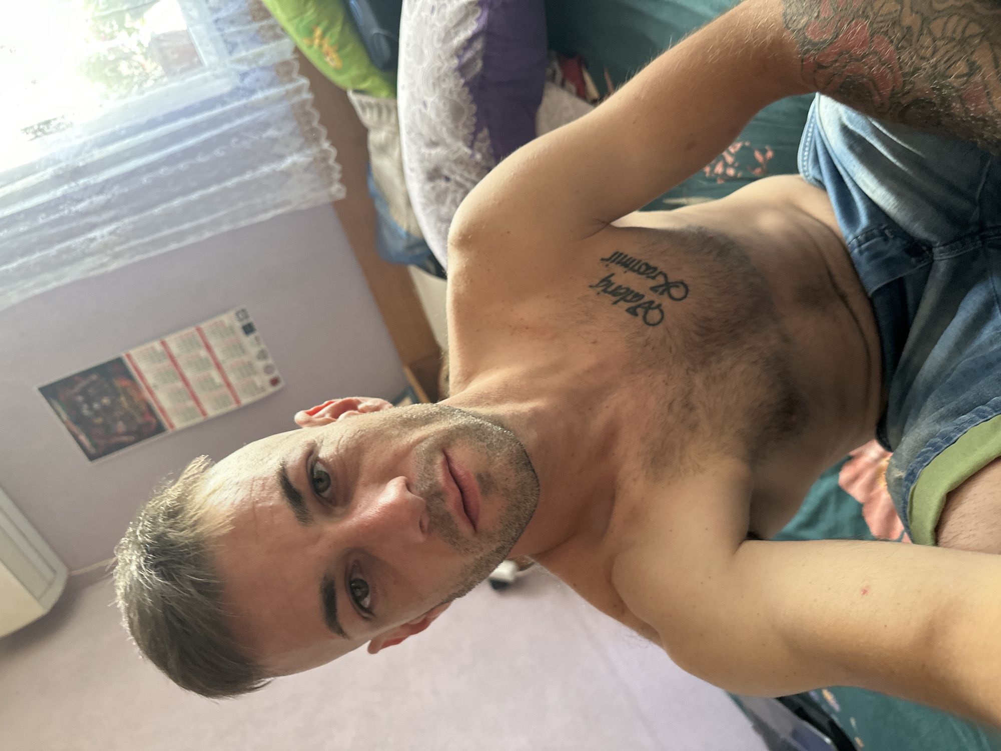 A young adult male is pictured in a medium shot, looking directly at the camera. He has a short, buzzed haircut with the sides shaved, and a light stubble on his face. His eyes are green, and he has prominent eyebrows. He is shirtless, revealing a tattoo on his chest that reads "Valeri Krasimir". He is wearing blue denim shorts. The background is a bedroom, with a wall, a window with white lace curtains, an air conditioner unit mounted high on the wall, and pillows on a bed. The overall lighting suggests it is daytime. There is no discernible indication of the specific location within Cherven Breg, Bulgaria.