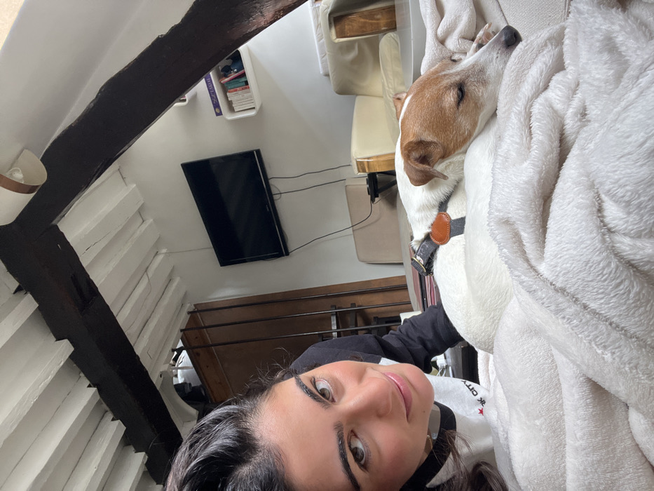 An interior scene depicts a female subject and a dog in a residential dwelling in Paris, France. The female, with dark hair, occupies the left foreground, looking directly at the camera with a slight smile. She wears a black and white top. To her right, a white and brown Jack Russell Terrier type dog is resting or sleeping on a white textured blanket, wearing a dark harness with a round, light-brown tag. In the background, a black flat-screen television is mounted on a white wall, below a white floating shelf containing books. Beige upholstered furniture is partially visible behind the dog. The ceiling features exposed dark wooden beams against white panels. A wooden cabinet or wardrobe is partially visible in the left background.
