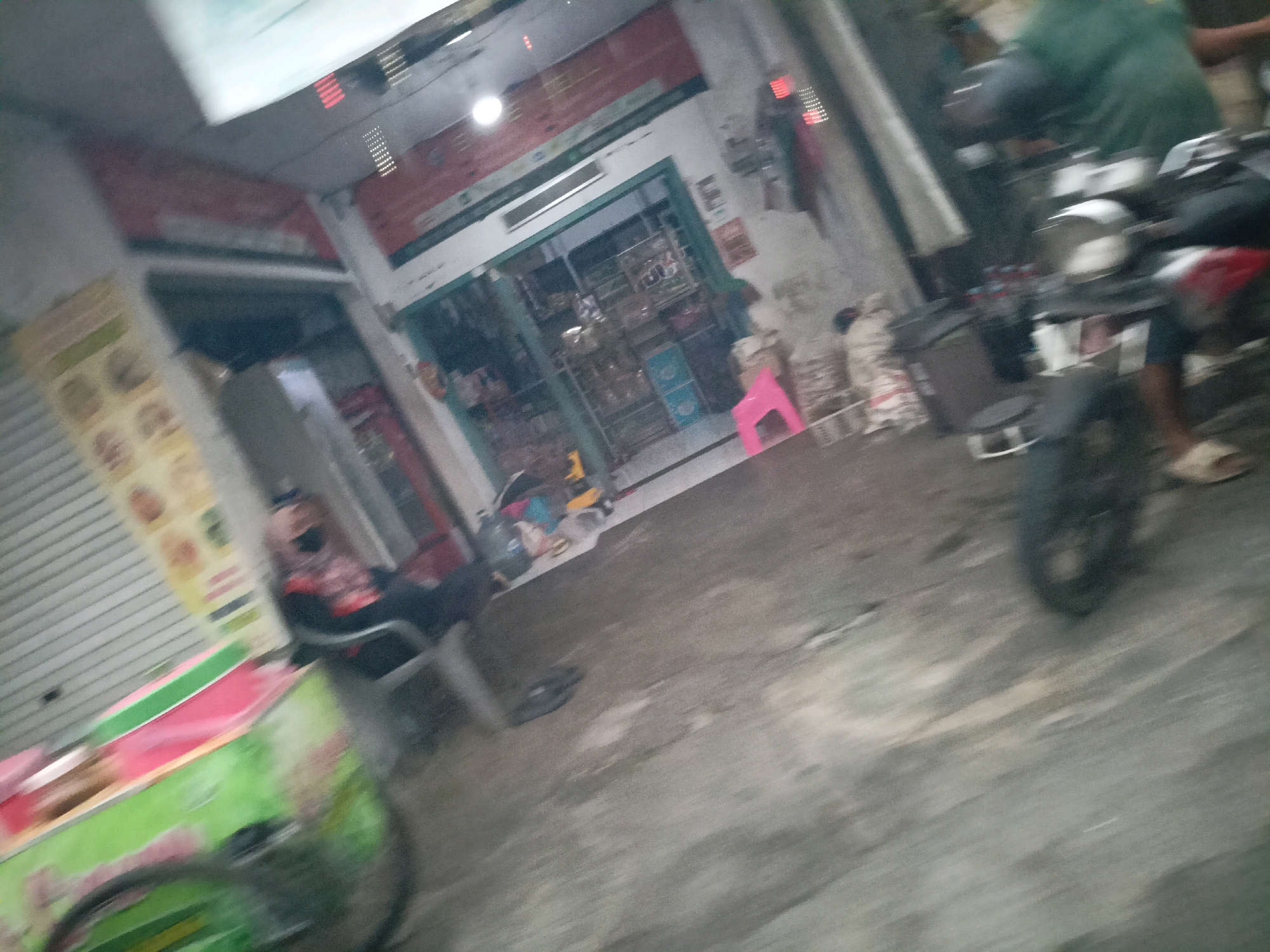 The image depicts a blurred outdoor scene in what appears to be a street or alleyway in Magelang, Indonesia. On the left, a person is seated in a chair, wearing a black mask and a patterned shirt, with their legs crossed. To the right of this person is a brightly colored cart with green and pink elements. In the background, a storefront is visible with shelves stocked with various items. A pink stool is placed in front of the store. Further to the right, a person wearing a green shirt is partially visible, and a motorcycle is present, with its rear wheel prominently featured. The ground is paved and appears wet in some areas. The lighting suggests it is either evening or nighttime.