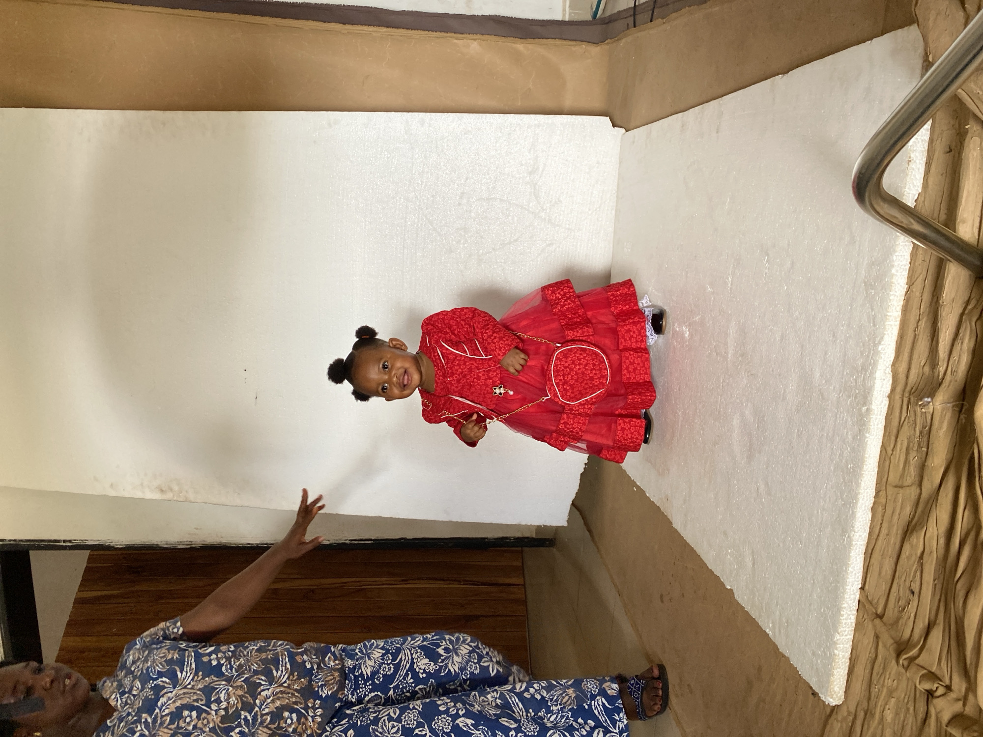 A young child in a red dress stands smiling near a woman in a blue floral outfit. The child is positioned in front of what appears to be a white foam board. The setting has brown walls and floors. The location is Bukuru, Nigeria.
