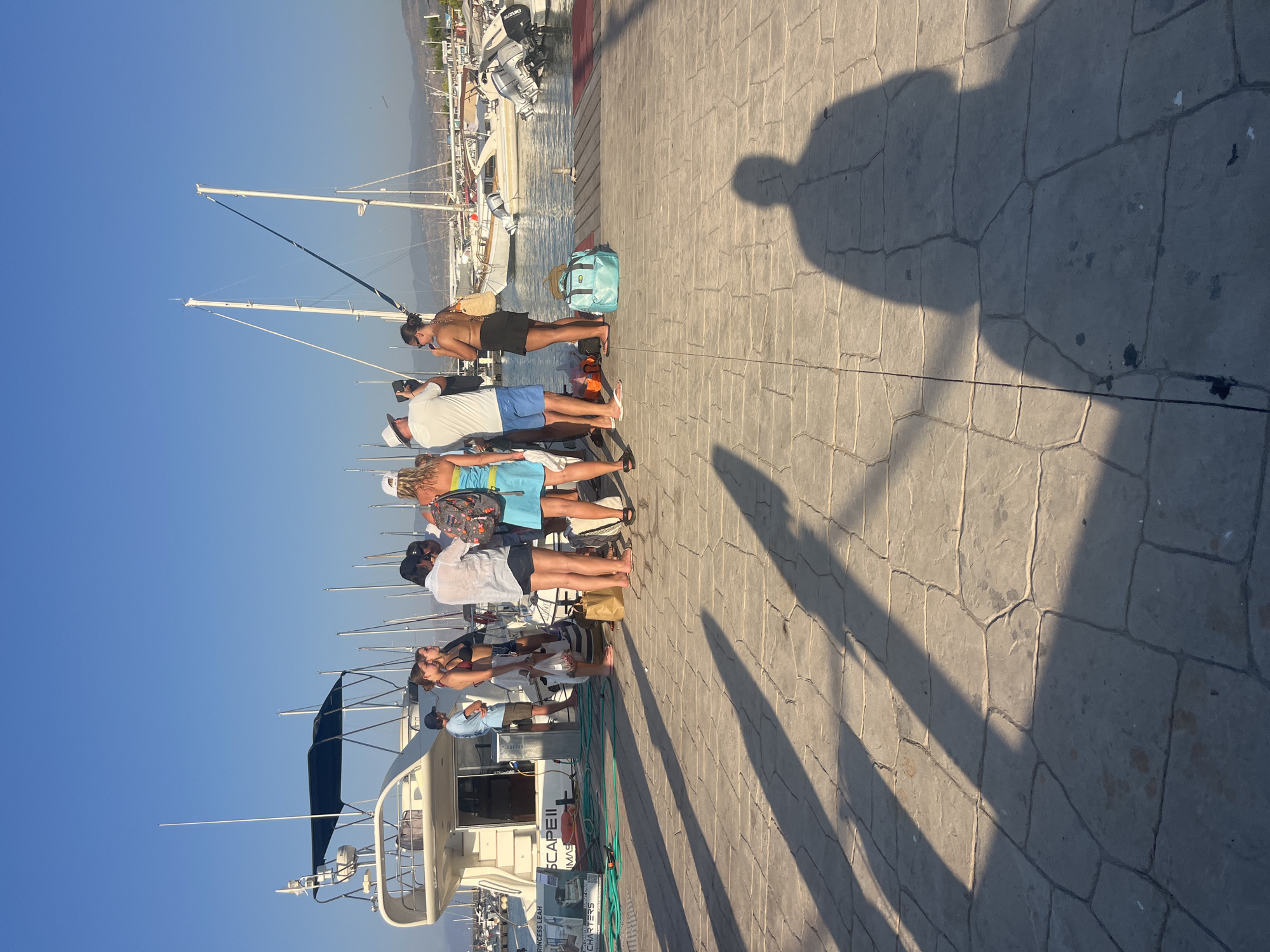 A group of seven people are standing on a stone-paved pier in Polis Chrysochous, Cyprus, near a white motor yacht named "PRINCESS LEAH ESCAPE II". The sky is clear and blue. The group consists of four women and three men, most of whom are dressed in casual summer clothing like swimsuits, shorts, and dresses. Some are holding bags, and one man appears to be taking a photo with his phone. Several sailboats and motorboats are docked in the background. The pier is illuminated by sunlight, casting long shadows on the stone surface.
