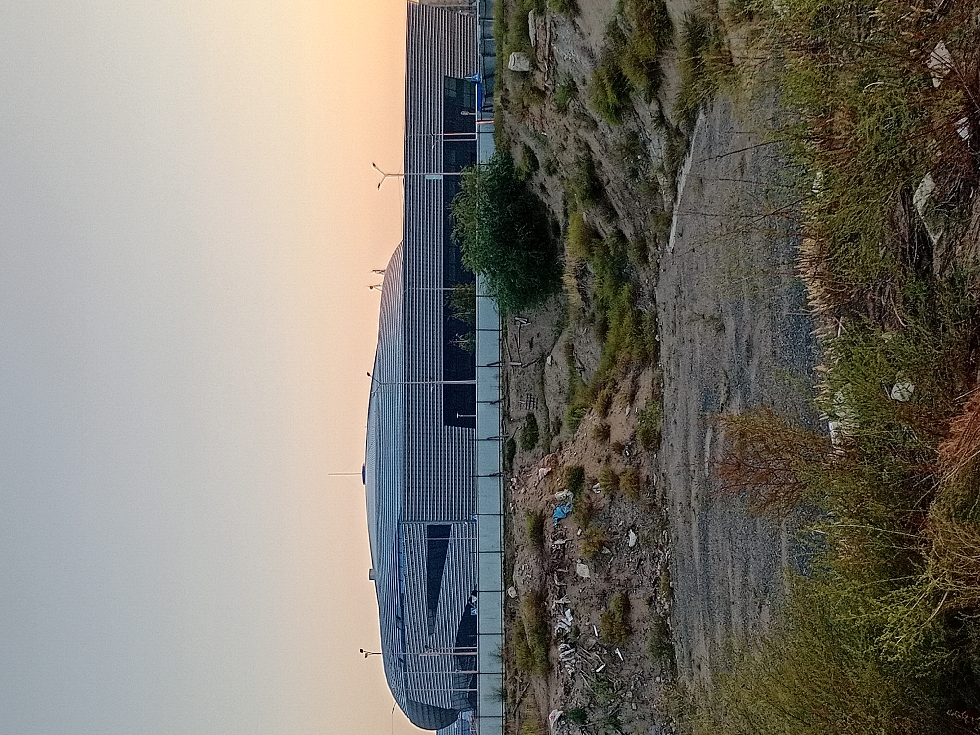 The image captures a large, modern building with a curved, metallic facade situated in Semey, Kazakhstan. The building, possibly a stadium or convention center, features horizontal louvers and reflective windows. It is set against a pale sky with hues of orange and yellow indicating either sunrise or sunset. The foreground and midground show a rough, unkempt landscape. There are mounds of dirt and debris, including what appears to be litter such as plastic bags and pieces of wood, scattered across the terrain. Sparse vegetation, including dry grasses and some green bushes, grows on these mounds. A low fence or barrier runs horizontally across the middle ground, separating the natural debris-filled area from the manicured grounds around the building. The overall impression is one of a juxtaposition between a contemporary architectural structure and a neglected, undeveloped surrounding environment. There are no people visible in the image.