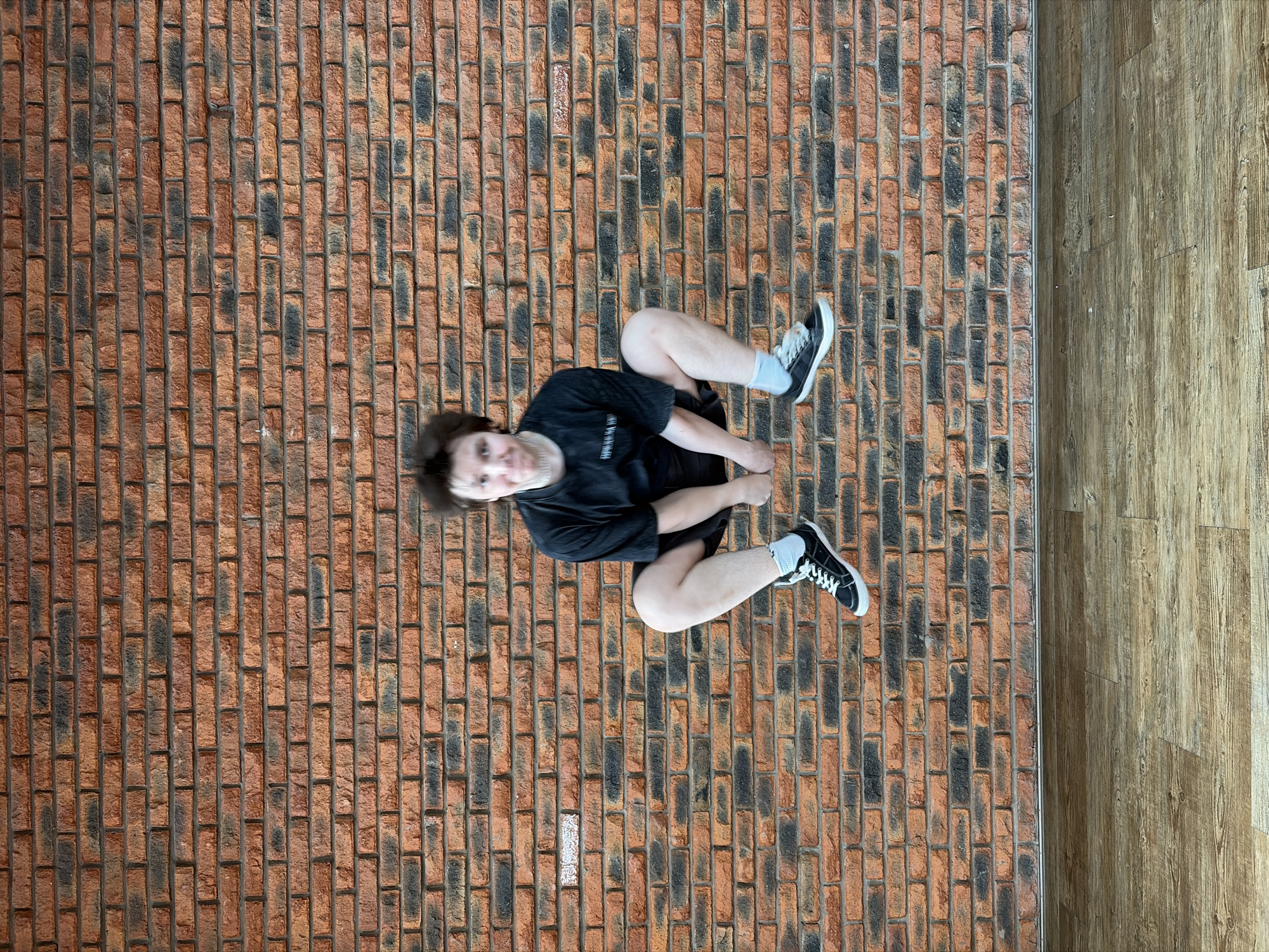 A person lies on a floor composed of two distinct surfaces: a patterned brick area and a wooden laminate section. The individual, wearing a black t-shirt, dark shorts, white socks, and black and white sneakers, is positioned such that their back rests on the brick section while their legs are bent. The person is facing towards the viewer and appears to be on the ground, the scene is likely to be indoors, but no other identifying features are visible in the image to suggest the location is in the city of Chaikovsky, Russia.