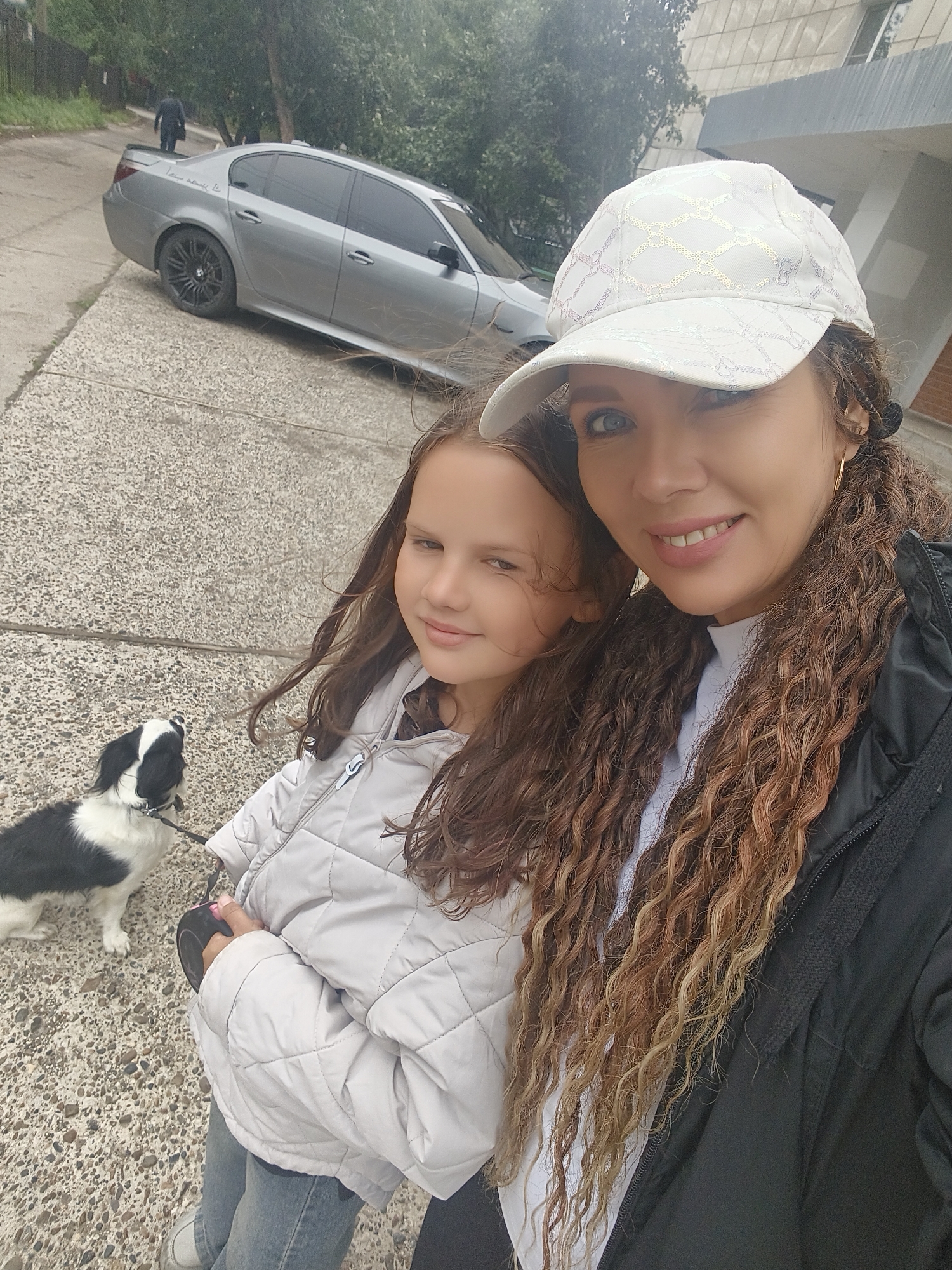 In the city of Unknown, Unknown, a woman and a young girl are walking a black and white dog. The woman is wearing a white cap and a black jacket, while the girl is wearing a light gray jacket. The dog is on a leash held by the girl. They are walking on a sidewalk next to a paved road. A silver BMW sedan is parked on the side of the road. Trees and a building are visible in the background.