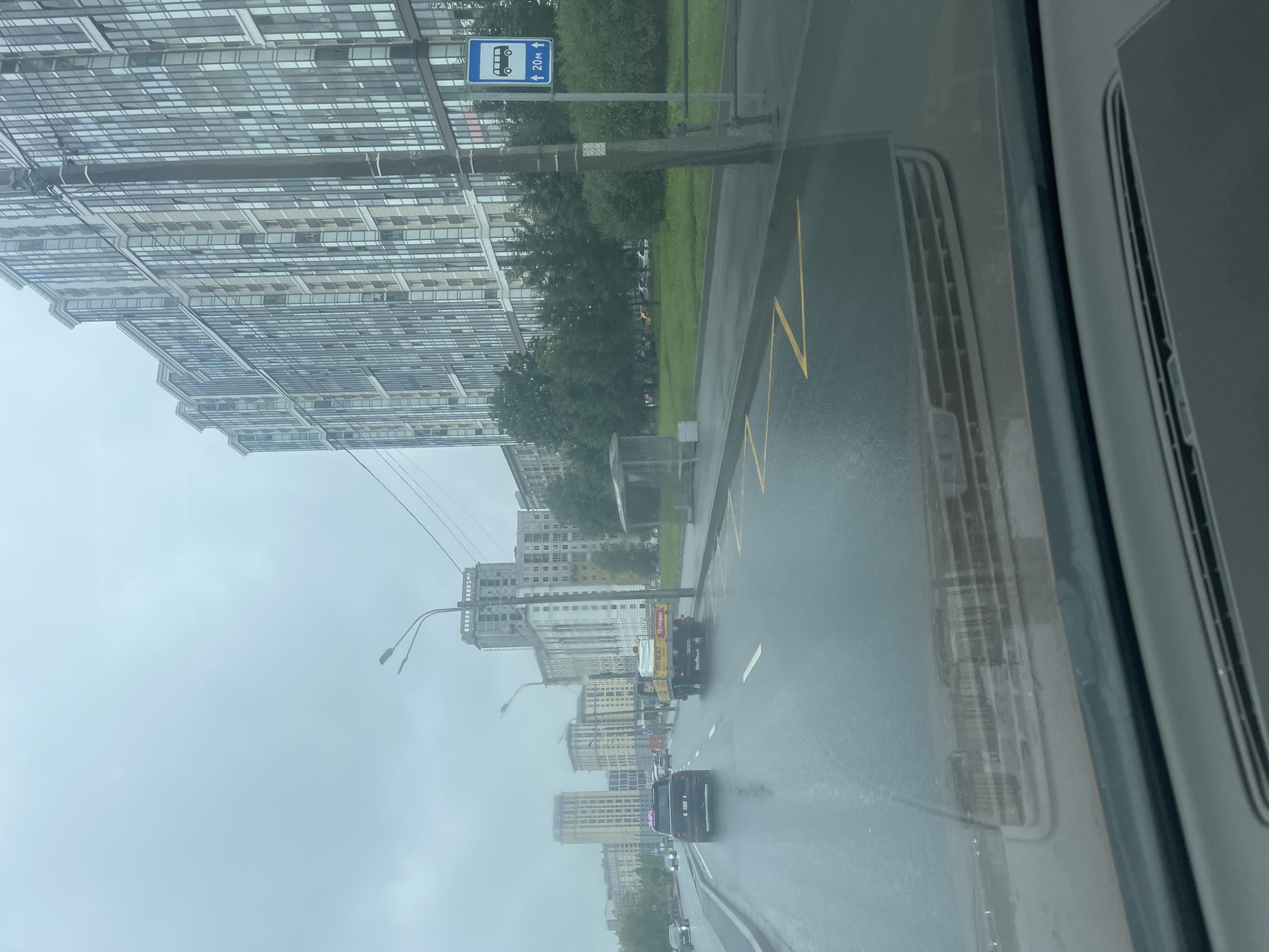 The image depicts a street scene in Saint Petersburg, Russia, likely in a modern residential district. The day is overcast, and the road is wet, indicating recent rain. The perspective is from inside a vehicle, looking forward through the windshield.
To the right of the road, a tall, contemporary apartment building with numerous windows dominates the background. Adjacent to the building, a line of trees forms a green buffer. A bus stop shelter with a blue sign indicating a bus stop 20 meters ahead is visible. A utility pole stands in front of the apartment building.
The wet asphalt road has yellow markings. Several vehicles are present on the road. A black SUV is in the left lane, and a flatbed truck carrying white materials is in the right lane. Other cars are visible in the distance, further down the road. In the background, other apartment buildings of varying heights and architectural styles are scattered across the city skyline. The sky is a uniform, dull gray. The foreground shows the interior of the vehicle, including the dashboard and windshield.
