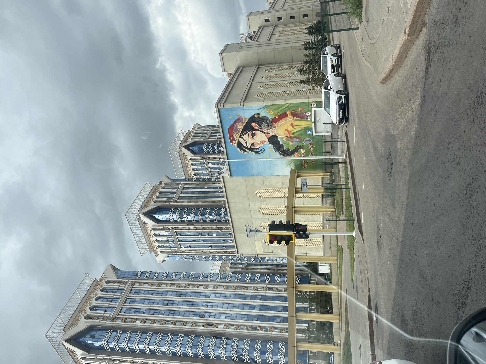 The image depicts a street view in Astana, Kazakhstan, featuring modern architecture and public art. Dominating the scene are several tall, sleek buildings with prominent blue windows and ornate decorative elements on their upper sections, indicative of contemporary urban design. A large mural of a young woman wearing traditional attire and a distinctive headdress is painted on the side of one of the buildings, adding a cultural touch to the urban landscape. The sky is overcast, with heavy clouds creating a diffused, somewhat dramatic lighting. A traffic light with a red signal is visible, suggesting the presence of regulated traffic flow. Two white cars are driving on the road in front of the muraled building, one in the foreground and another slightly behind it. The road is asphalt, with a sidewalk and some greenery visible along the side. The overall impression is one of a developed, modern city with a blend of contemporary architecture and culturally significant art.