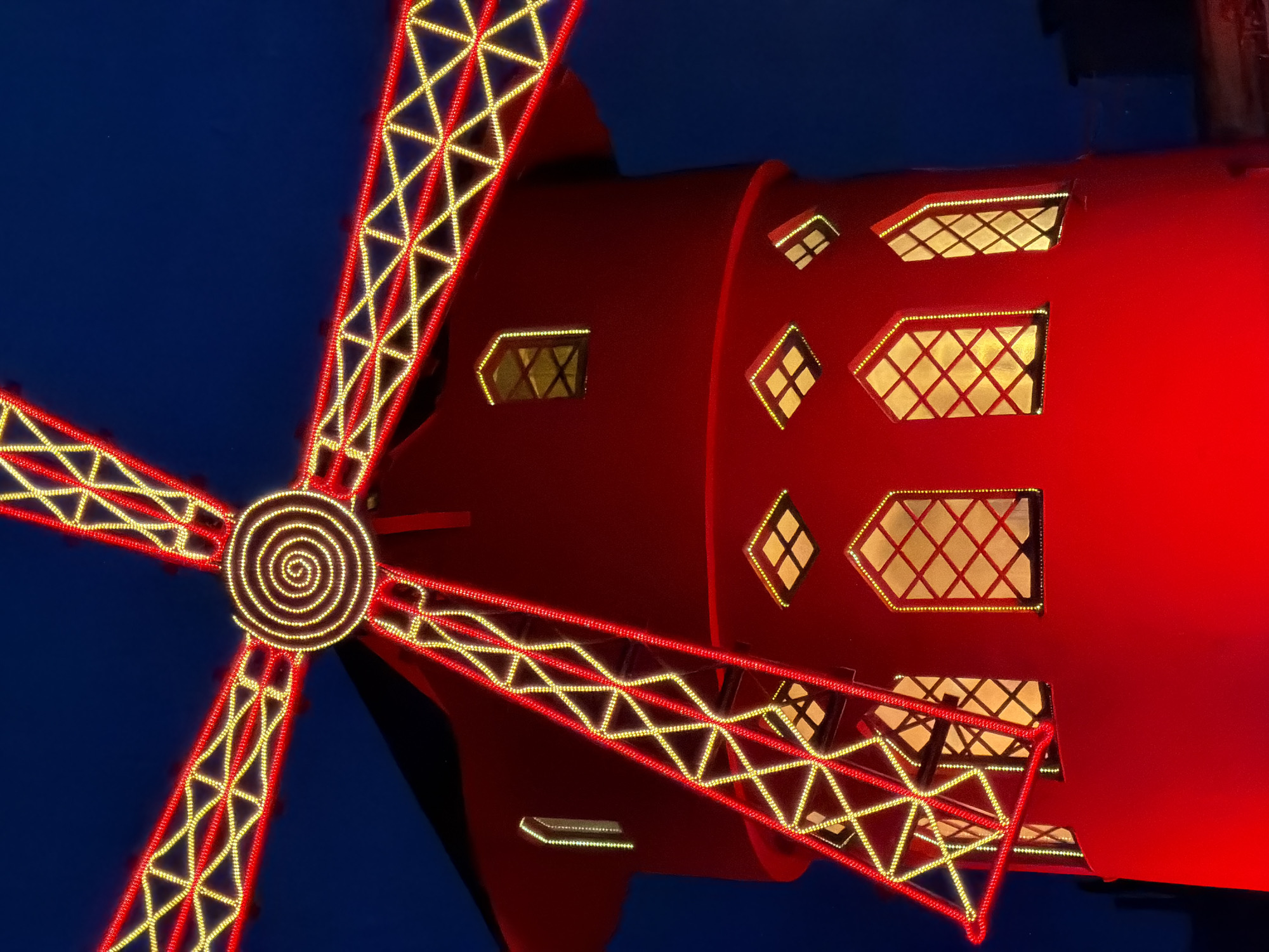 The image depicts the illuminated facade of the Moulin Rouge in Paris, France at night. The red windmill sails are decorated with yellow and red lights, forming a triangular latticework pattern. A spiral of lights adorns the center. The building itself is red, with various windows lit from within, some diamond-shaped and others rectangular with crisscrossed patterns. The background is a dark blue, suggesting the night sky.