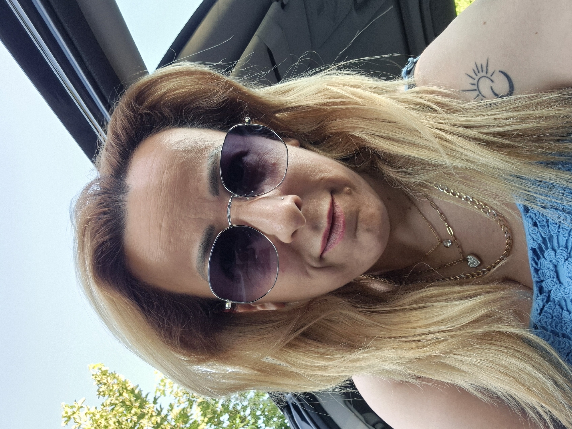 A medium close-up, selfie-style shot shows a woman with blonde hair and sunglasses. She is outdoors, with a blue sky and green foliage visible in the background. The woman is wearing a blue top and several necklaces, including one with a heart-shaped pendant. A tattoo of a sun and crescent moon is visible on her left shoulder. She appears to be in the driver's seat of a car, as indicated by the sunroof and car interior visible in the background. The image is well-lit and appears to have been taken during the day.