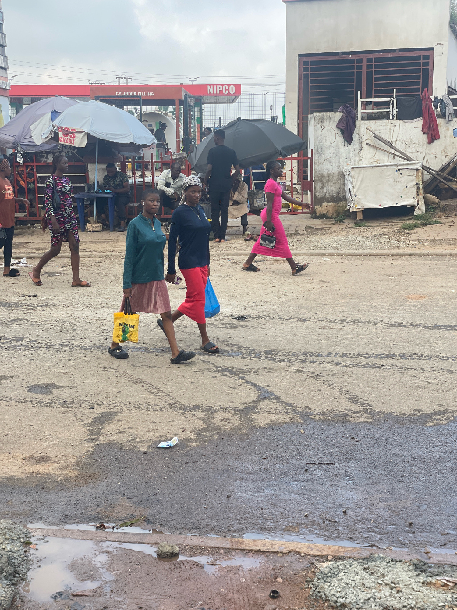 The image depicts a street scene in Abuja, Nigeria, with a gas station in the background. The foreground features a paved road with visible tire marks and puddles of water, indicating recent rain. Several people are walking across the road. Two women are in the center of the frame, walking side-by-side. The woman on the left wears a teal long-sleeved top and a pink pleated skirt, carrying a yellow bag. The woman on the right wears a navy blue long-sleeved top and a red skirt, holding a blue plastic bag. To the right of them, a woman in a bright pink dress walks away from the camera, carrying a small black purse. Further to the left, another woman in a patterned dress is walking towards the foreground. In the background, a gas station with the "NIPCO" logo and "CYLINDER FILLING" signage is visible, along with several covered stalls and a building with a corrugated metal roof. People are seen gathered at the gas station and under the stalls. The sky is overcast.