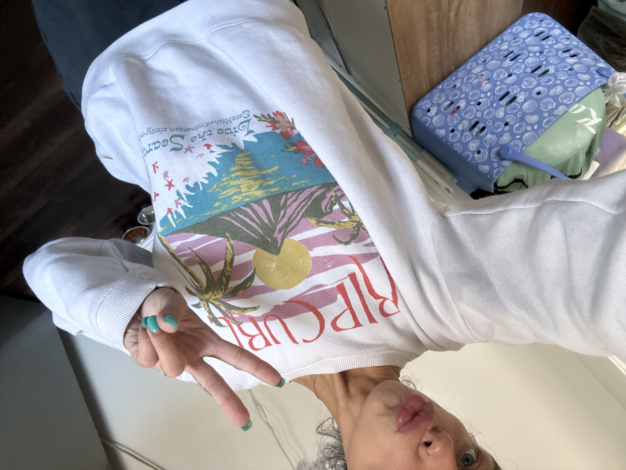 A woman is posing for a selfie, wearing a white sweatshirt with "RIP CURL" printed on the front with a beach scene. She is making a peace sign with her left hand, which has green nail polish. Her face is visible, showing her nose and lips. A blue plastic basket with a white seashell pattern is in the background, as well as a wooden table and a wall. The location in the city of Seal Beach, United States of America, cannot be determined from the available information.