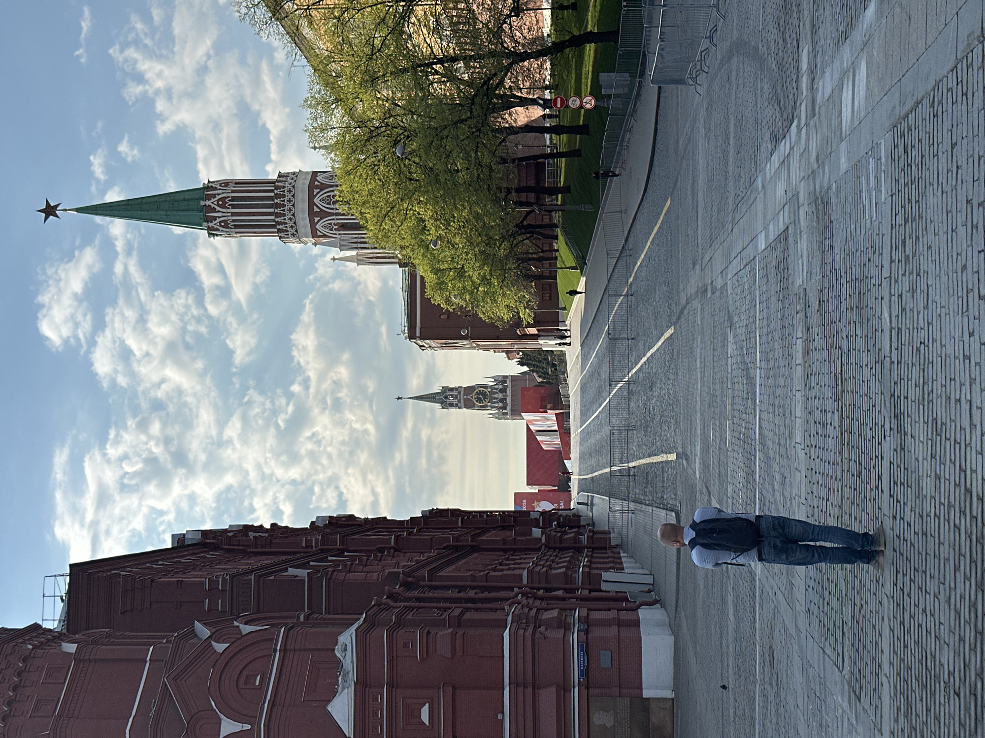 The image shows a street scene in Moscow, Russia. Prominently featured are the towers of the Moscow Kremlin, specifically the Spasskaya Tower and another tower with a green roof and red star at the top. A red brick building, part of the Kremlin wall, is visible on the left side of the frame. The ground is paved with cobblestones, and a man wearing jeans and a backpack is walking away from the viewer on the right side of the street. A grassy area with trees is present on the other side of the Kremlin wall, and a blue sky with scattered clouds forms the backdrop.