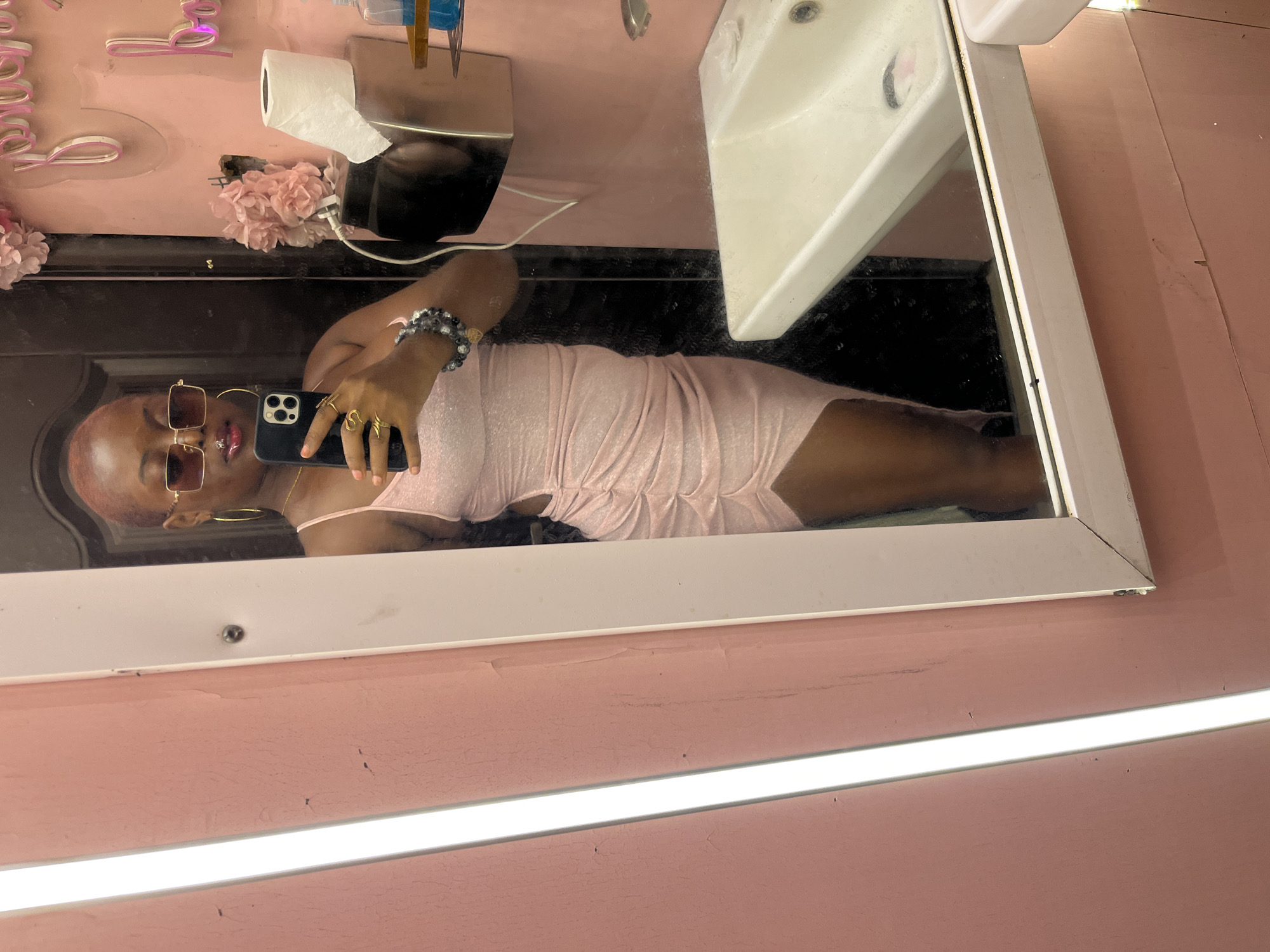 A full-length mirror reflects a young Black woman taking a selfie. She has short, reddish-pink hair, wears large gold hoop earrings, gold-rimmed square sunglasses, and a pink, sparkly, ruched bodycon dress with cutouts on the sides. She is also wearing a silver ring and a beaded bracelet. She is holding a black smartphone in her right hand, using it to take the photo. The background is a pink-walled room, possibly a bathroom. To the right of the mirror, a white sink is visible, along with a roll of toilet paper and a dispenser of hand sanitizer. Decorative pink flowers and neon pink script lettering are affixed to the wall behind the mirror. The overall aesthetic is bright, feminine, and possibly indicative of a salon or boutique. The location is stated to be Howayi, Nigeria.