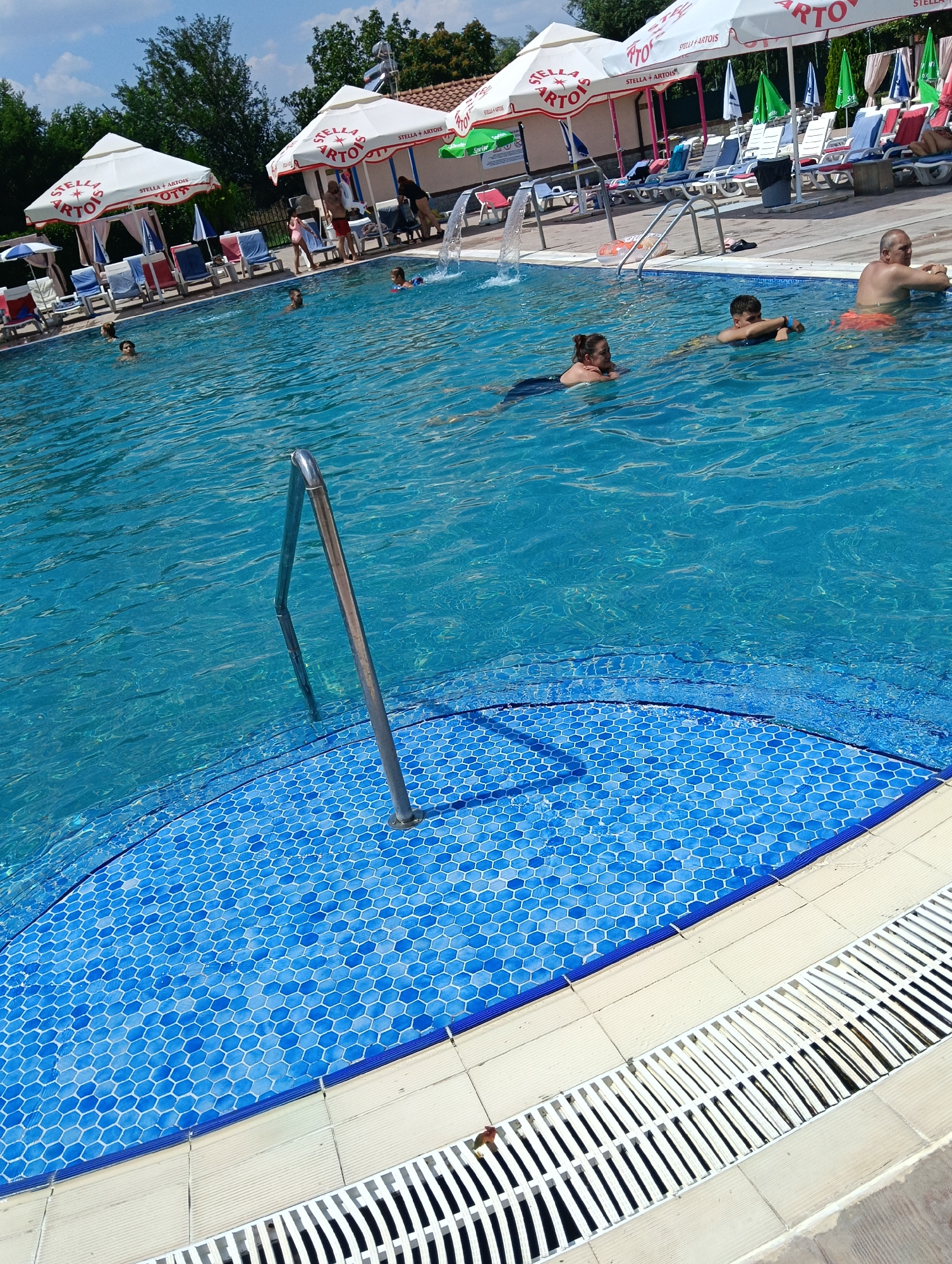 This is a poolside scene at a public swimming pool located in Radievo, Bulgaria. The pool is filled with clear blue water and has a tiled bottom. Several people are visible in the water, including adults and children, engaged in swimming and leisure activities. Surrounding the pool are lounge chairs and umbrellas, suggesting a recreational area. The overall environment is sunny and appears to be a warm day.