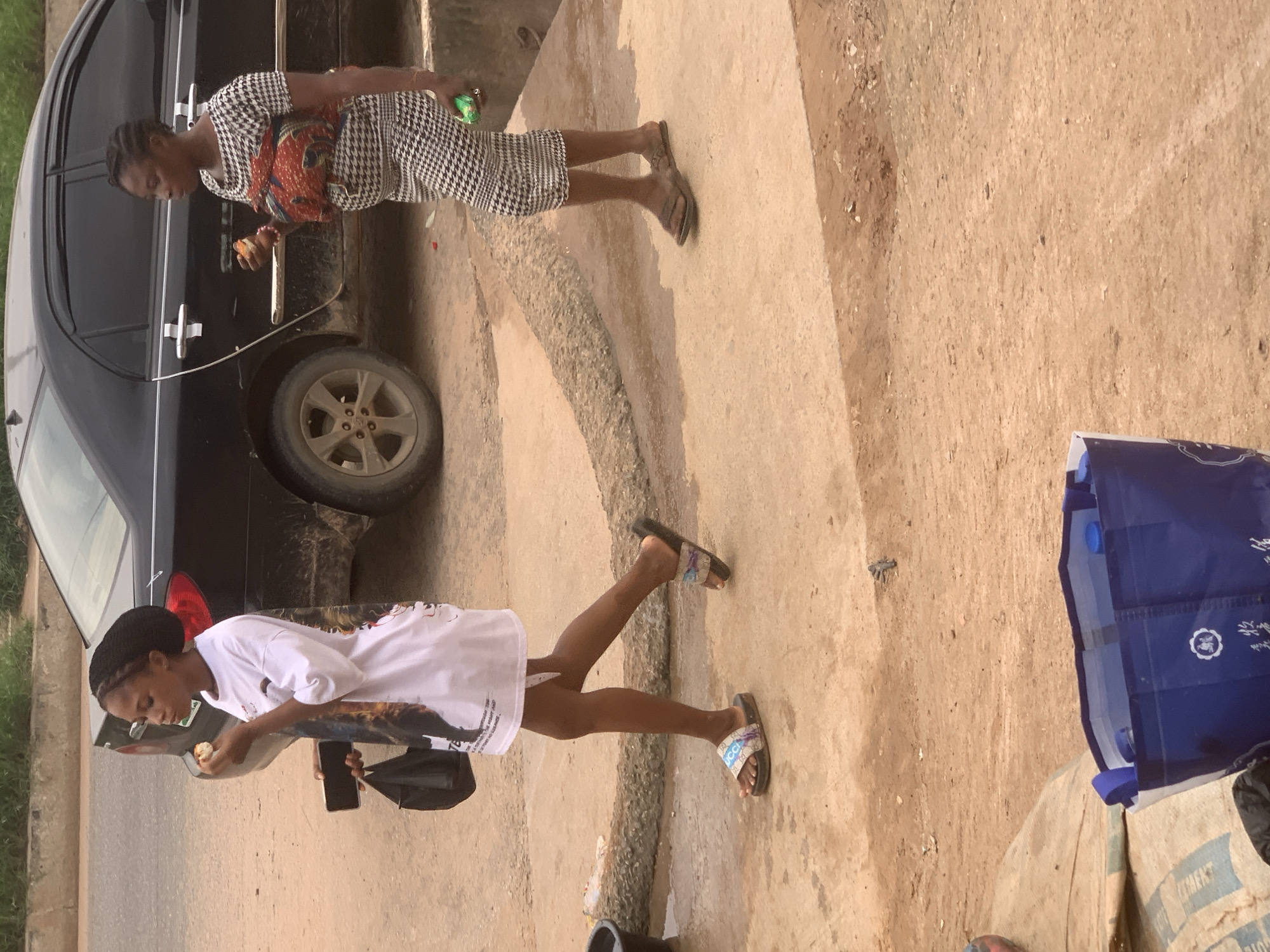 In Shagamu, Nigeria, two women are standing near a black sedan car parked on a dirt road. The woman on the left, with a black braided hairstyle, is wearing a white t-shirt with a graphic print and a black bag over her left shoulder. She is holding a smartphone in her left hand and eating a piece of food with her right. The woman on the right is wearing a black and white checkered dress and has a baby wrapped in a patterned fabric on her chest. She is holding a green object in her right hand and also appears to be eating. The environment is dusty and unpaved, with some vegetation visible in the background. A blue tote bag is on the ground in the foreground.
