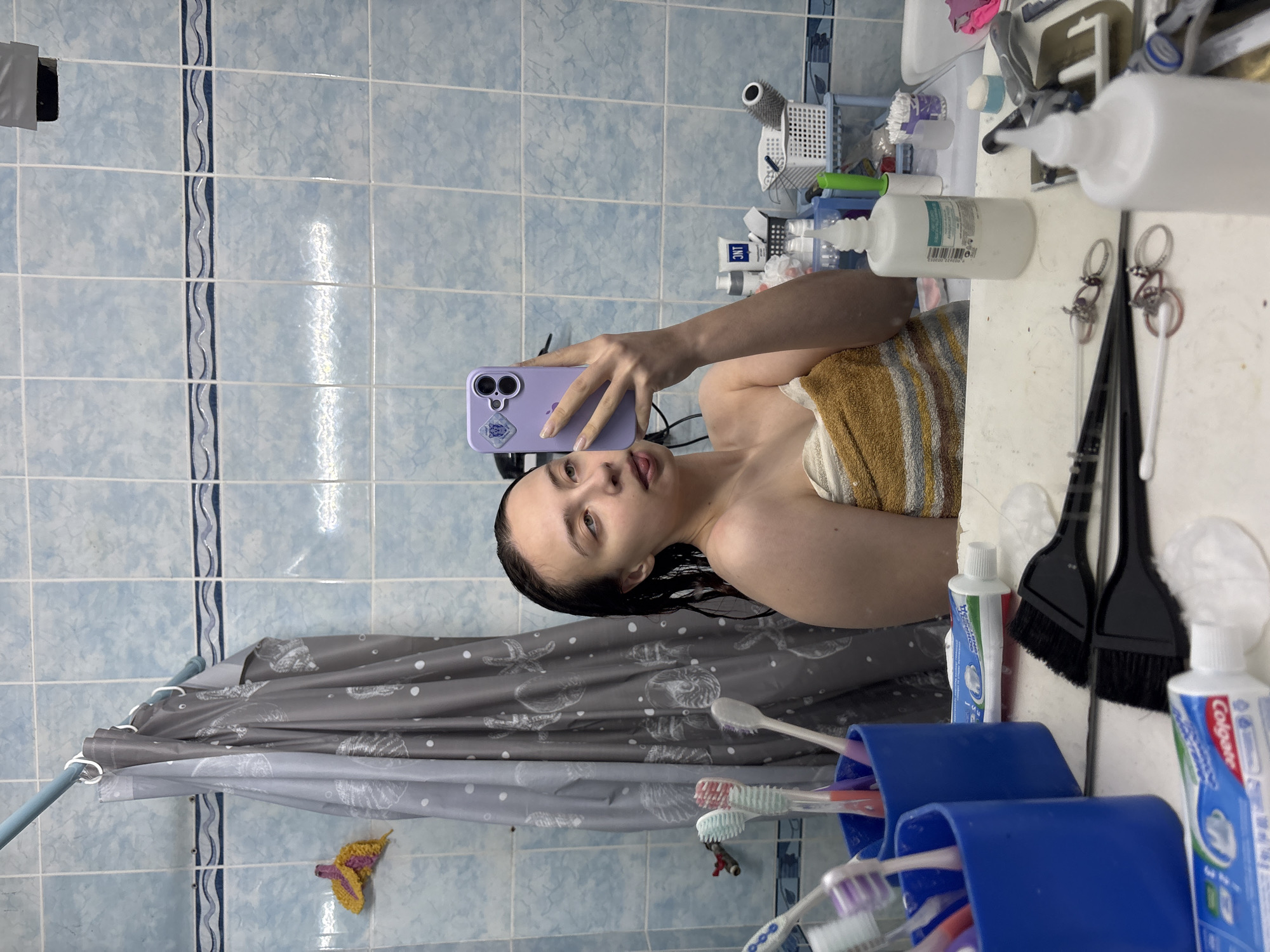 The image shows a young woman taking a selfie in a bathroom. She is holding a purple smartphone with a sticker on its back, and she is wrapped in a striped towel. Her hair appears to be wet.
The bathroom has blue tiled walls with a decorative blue-and-gray vertical stripe. A shower curtain with a marine-themed pattern (starfish and shells) is visible in the background, along with a crocheted yellow butterfly.
The foreground includes a mirror reflecting the scene. On the vanity or shelf near the mirror are various toiletries: toothpaste tubes (Colgate, Lacalut), black makeup brushes, and plastic containers with toothbrushes.
Based on the available information, the location is most likely a bathroom within the city of Прикамский, Russia.