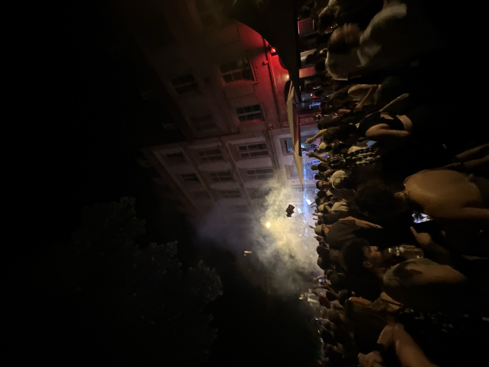 The image depicts a nighttime outdoor gathering of a large crowd of people, possibly a festival or street party, in Strasbourg, France. The scene is illuminated by ambient light from buildings and stage lighting, with visible smoke or fog machines creating a hazy atmosphere. Several individuals are captured in motion, with hands raised, suggesting a lively and energetic environment. In the background, multi-story buildings with lit windows line a street, typical of an urban European setting. One building in the middle distance is prominently lit by red lights. The foreground is densely populated with people, many of whom are silhouetted against the light sources. The overall impression is of a vibrant social event taking place at night in a city setting.