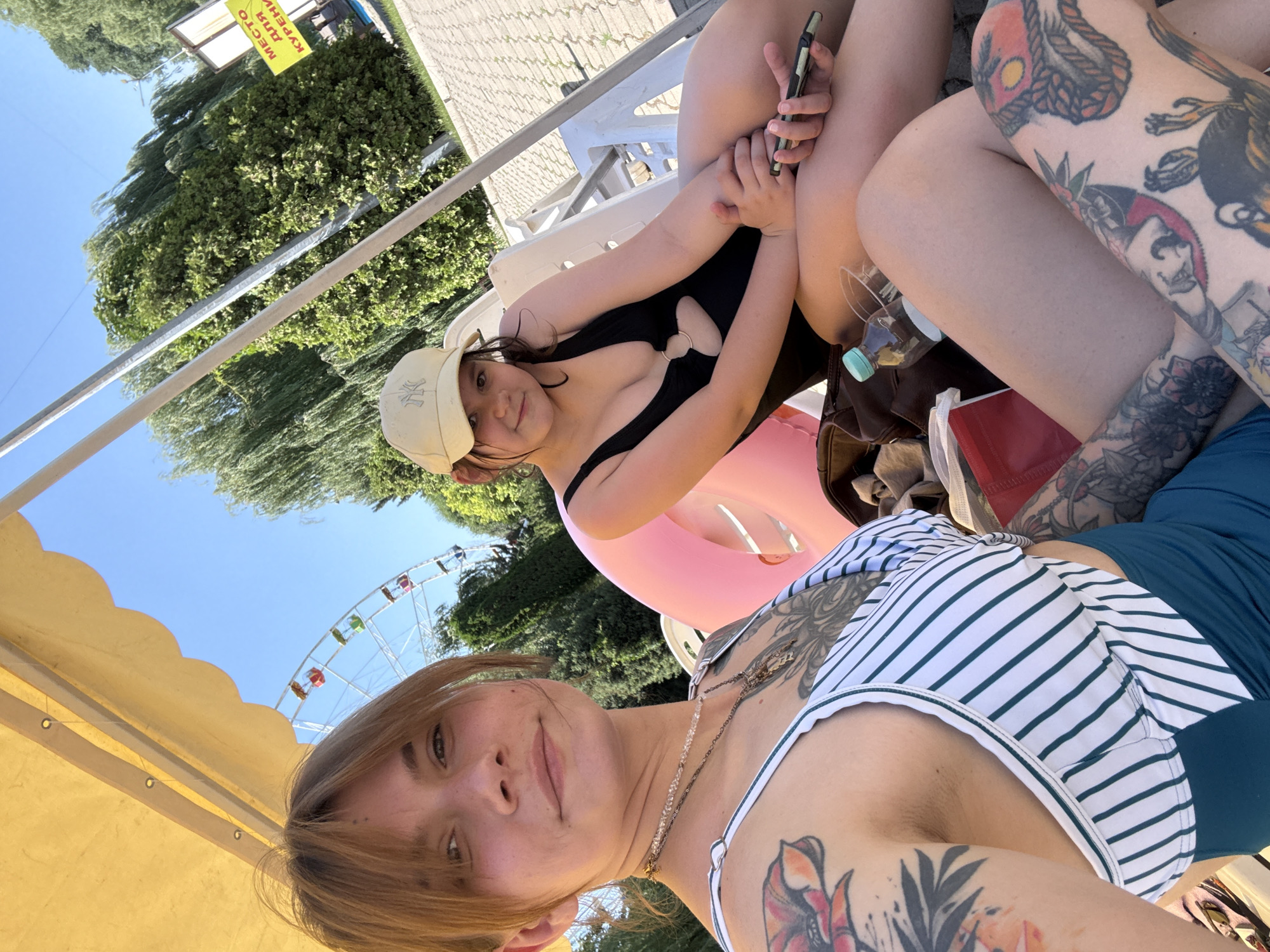 The media file shows two young women outdoors on a sunny day. One woman in the foreground is wearing a striped bikini top and has blonde hair and tattoos. The other woman, partially visible in the background, is wearing a black swimsuit and a light-colored baseball cap, and also has tattoos. They appear to be at a poolside or a similar recreational area, possibly an amusement park, as a Ferris wheel is visible in the background against a clear blue sky. There are trees and lounge chairs around them, suggesting a warm and relaxed atmosphere. One woman is holding a smartphone. A yellow sign with Cyrillic text is visible on the left side of the image, which reads "МЕСТО ДЛЯ КУРЕНИЯ" which translates to "SMOKING AREA". The image seems to be taken during daylight hours.