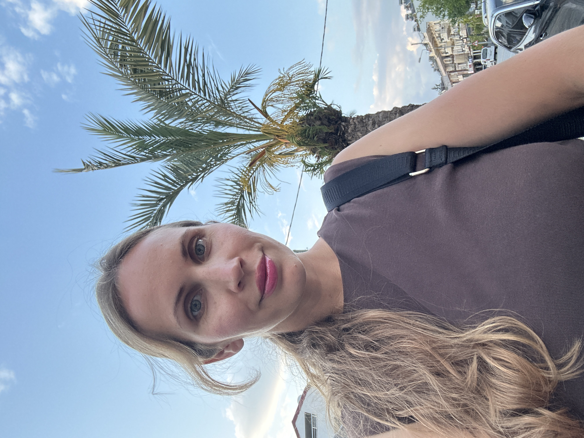 A selfie taken in Fethiye, Turkey, features a blonde woman with light eyes wearing a brown t-shirt and a black shoulder bag. A palm tree is visible in the background, along with buildings and a vehicle parked on the street. The sky is blue with scattered clouds.
