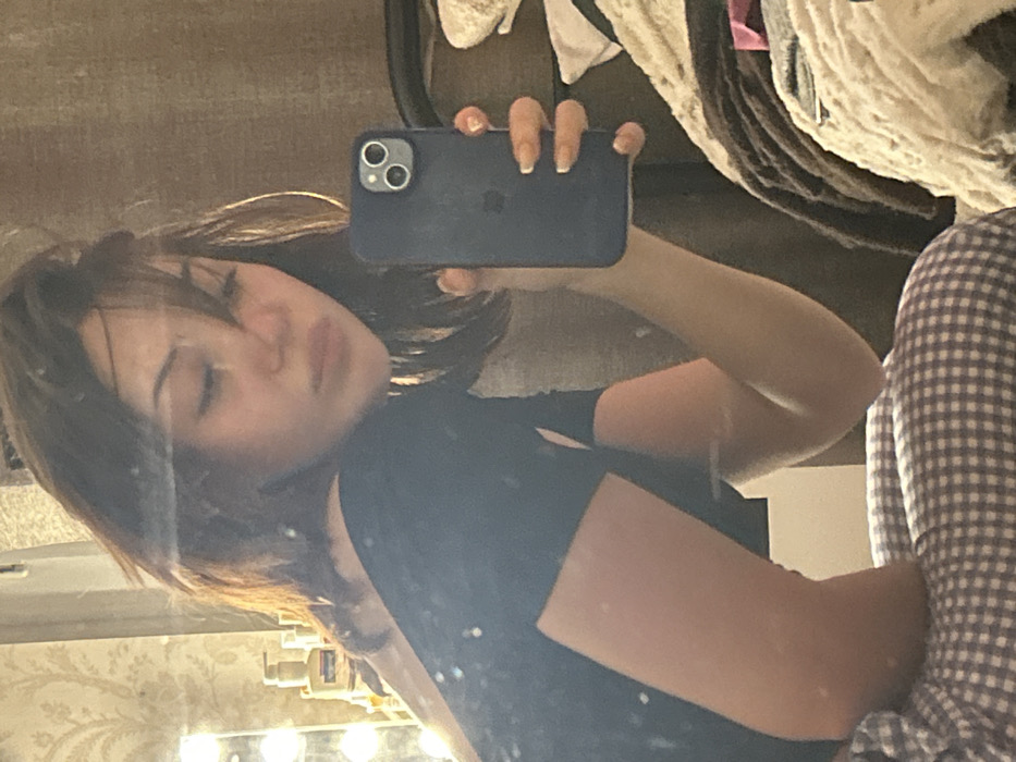 A selfie is taken in a mirror by a woman holding a dark blue iPhone. Her face is slightly blurred with her eyes partially closed. She wears a black short-sleeved shirt. Clutter is visible behind her, including blankets and a patterned shirt. The mirror reflects a vanity area with lights and various bottles. Her nails are long and painted a light color. The overall composition is somewhat unfocused and reflects a casual, candid moment.