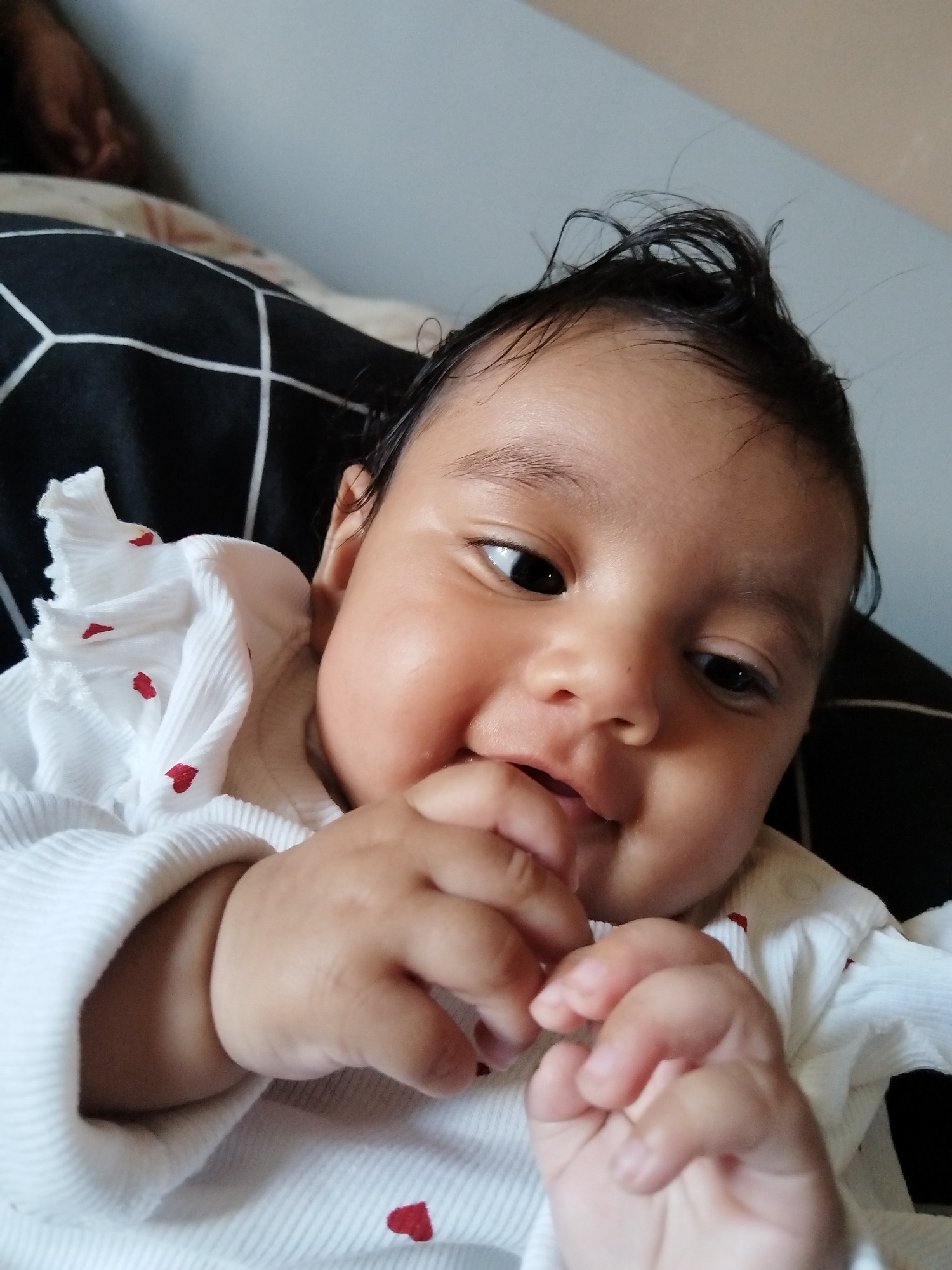 The image shows a close-up of a baby lying down. The baby has dark hair and eyes, and is wearing a white shirt with red hearts. The baby is holding their hands near their mouth and appears to be smiling or smirking. The background includes a black and white patterned surface. The location is Unknown, Unknown.
