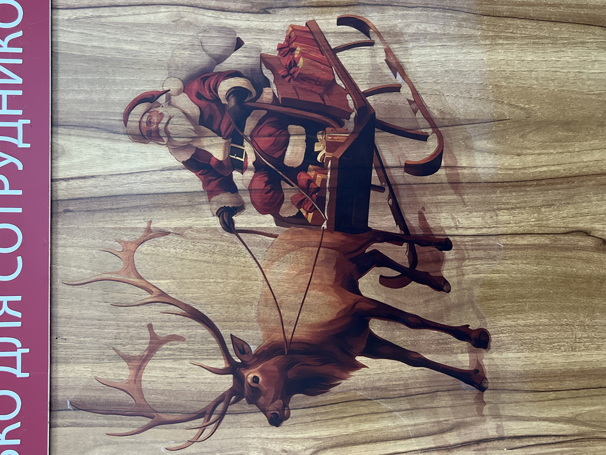 In Kadamjay, Kyrgyzstan, a decorative panel features a depiction of Santa Claus riding in a sleigh pulled by a reindeer. The panel is made of wood with a visible grain texture. Santa Claus, wearing his traditional red suit and hat, is seated in a dark-colored wooden sleigh laden with red gift boxes. The reindeer, with large antlers, is positioned to the left of Santa, pulling the sleigh. The background of the image is a light brown wood grain. At the top of the image, a red banner with light blue Cyrillic lettering is visible. The scene appears to be an outdoor decoration or advertisement, likely related to a winter holiday theme.