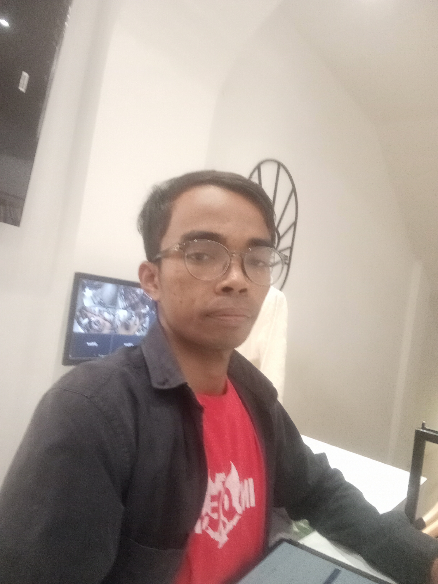 A man wearing glasses, a dark-colored shirt over a red t-shirt, and a dark-colored jacket is pictured in what appears to be a retail store in Larangan, Indonesia. The man is looking forward, and his right arm is resting on a table where a tablet is visible. Behind him, a security monitor displays multiple camera feeds, and a decorative circular metal object is mounted on the wall. The lighting is bright, suggesting an indoor setting.