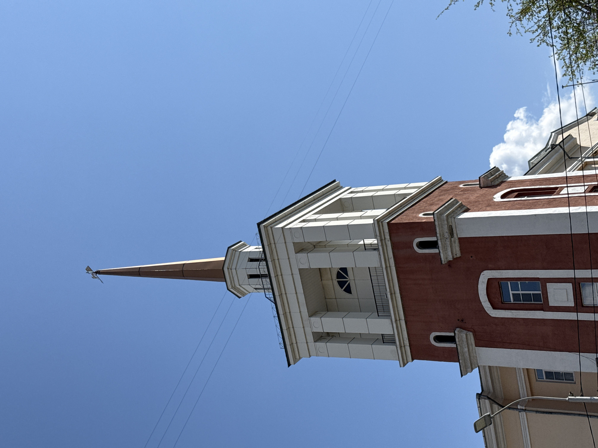 The image shows a tower with a spire, likely located in Krasnoyarsk, Russia, based on architectural styles found in the city. The tower is red and white with a multi-tiered design, featuring balconies and decorative columns. A long, pointed spire extends from the top of the tower. The sky is clear and blue. Electrical wires run in front of the tower. Parts of adjacent buildings and foliage are visible.
