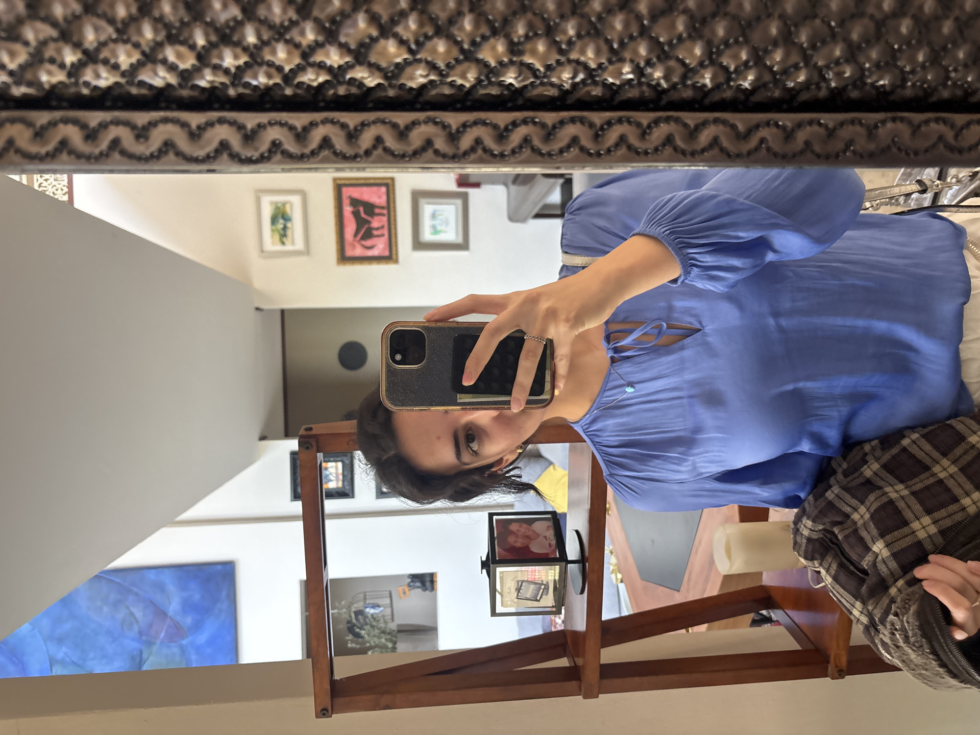 A person is taking a selfie in a mirror. The mirror has an ornate dark frame. The person is wearing a blue top. Reflected in the mirror are various framed artworks on the wall. A wooden stand with rotating picture frames is also visible. A candle and a plaid jacket sit on a surface, likely a table. While the location in Puebla, Mexico, cannot be confirmed from the image, the architectural style and artwork could be indicative of such a location.